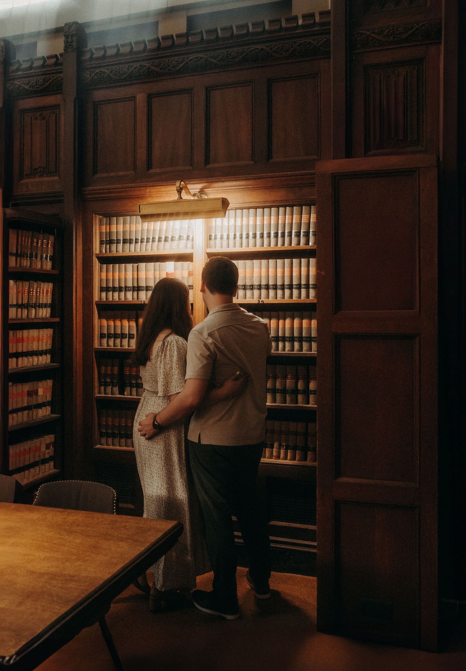 A couple browses books together in a warm-lit library with vintage wooden shelves and classic leather-bound volumes. Engagement Photography