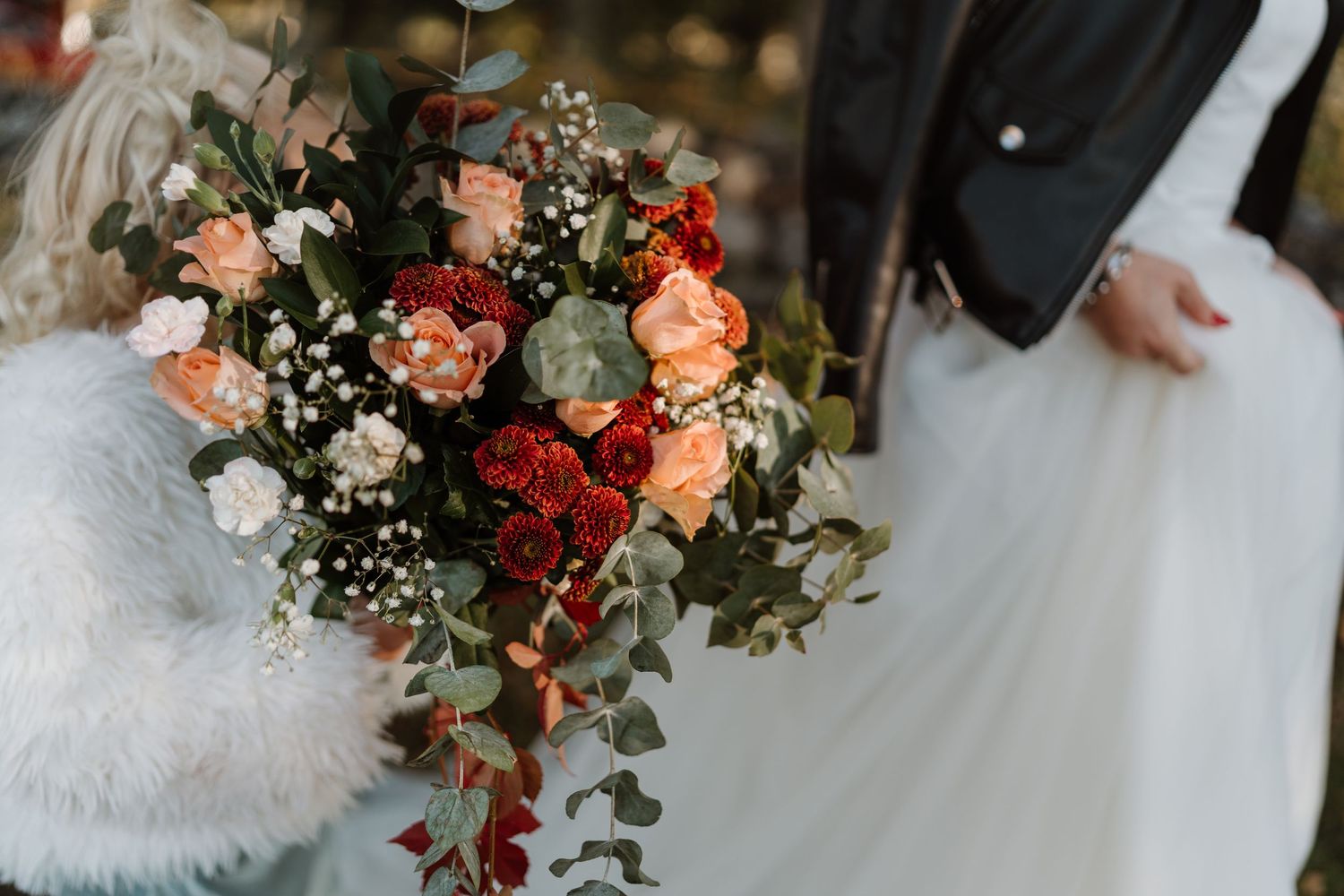 Bride holding a lush bouquet of peach roses, red dahlias, and eucalyptus at a wedding ceremony.
