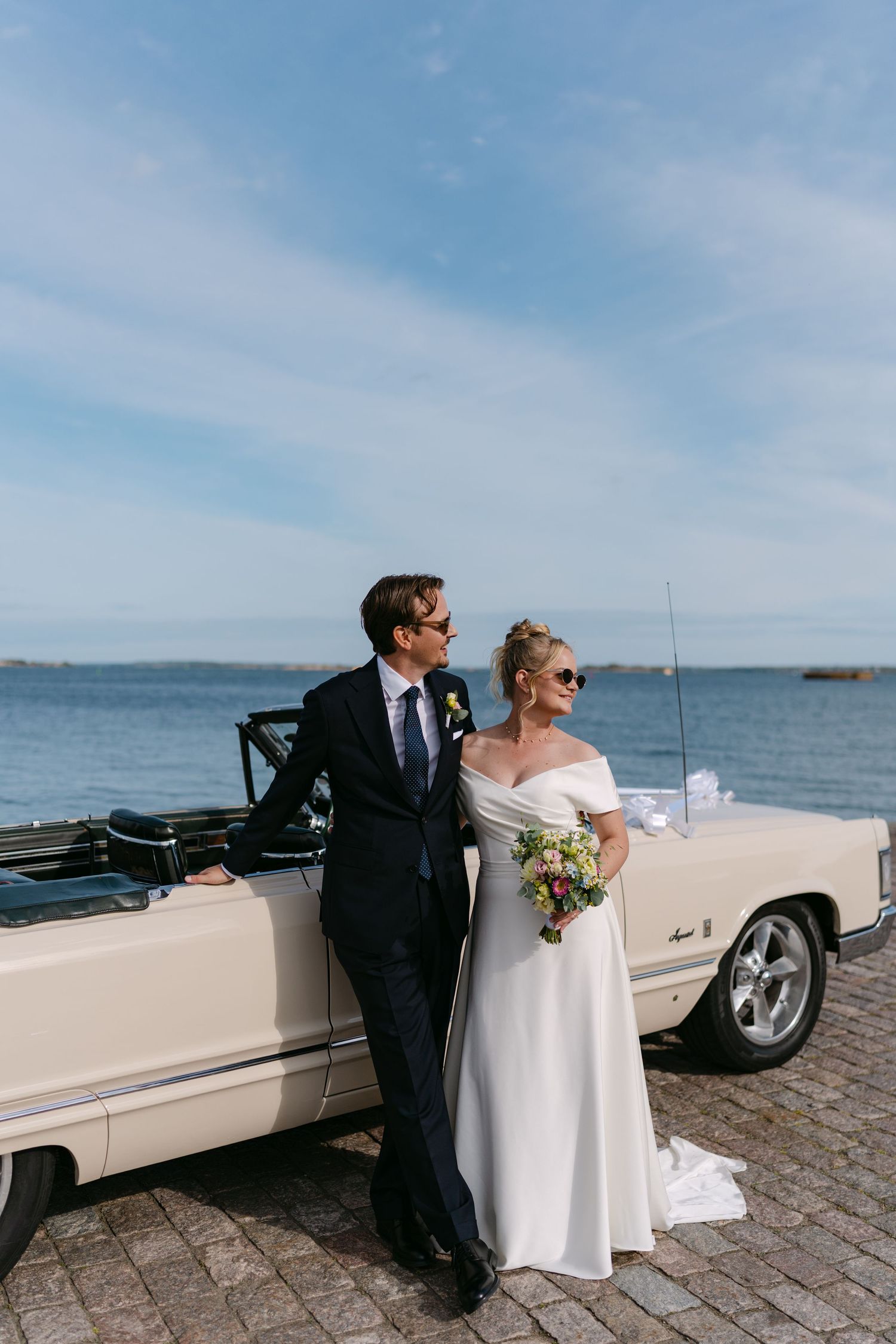 Bride and groom posing by a vintage cream convertible car near the ocean on their wedding day.