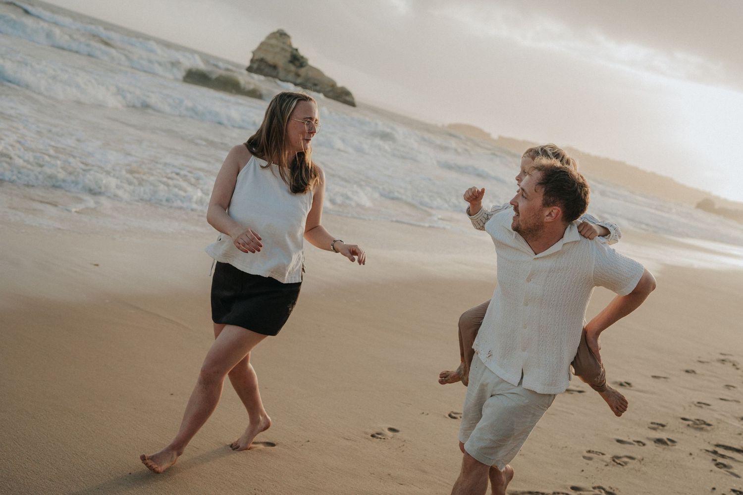 Couple laughing and playing on a sandy beach at sunset with ocean waves in the background.