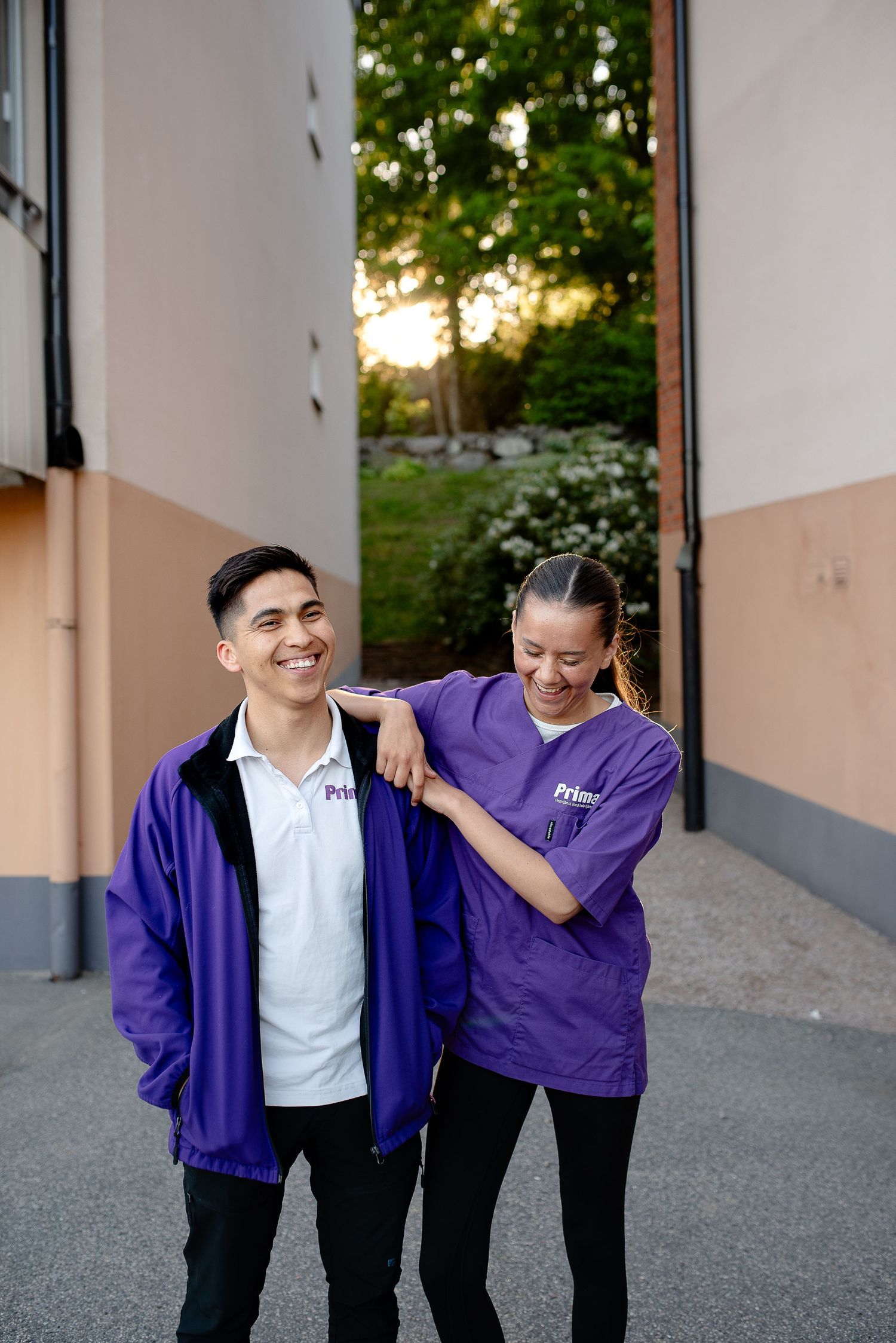Two smiling coworkers in purple branded uniforms standing together outdoors near a building entrance.