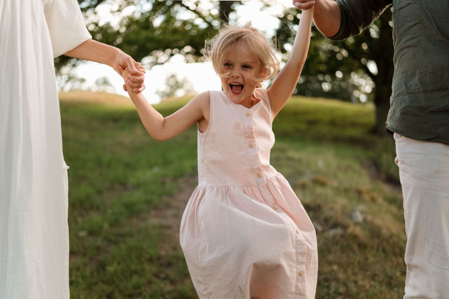 Joyful toddler girl in pink dress laughing while being swung by parents hands outdoors in green field.