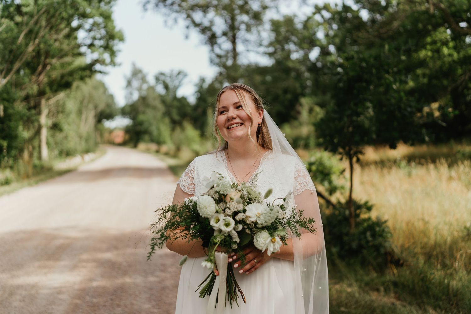 A bride laughs joyfully while holding a lush white and green bouquet on a country road.