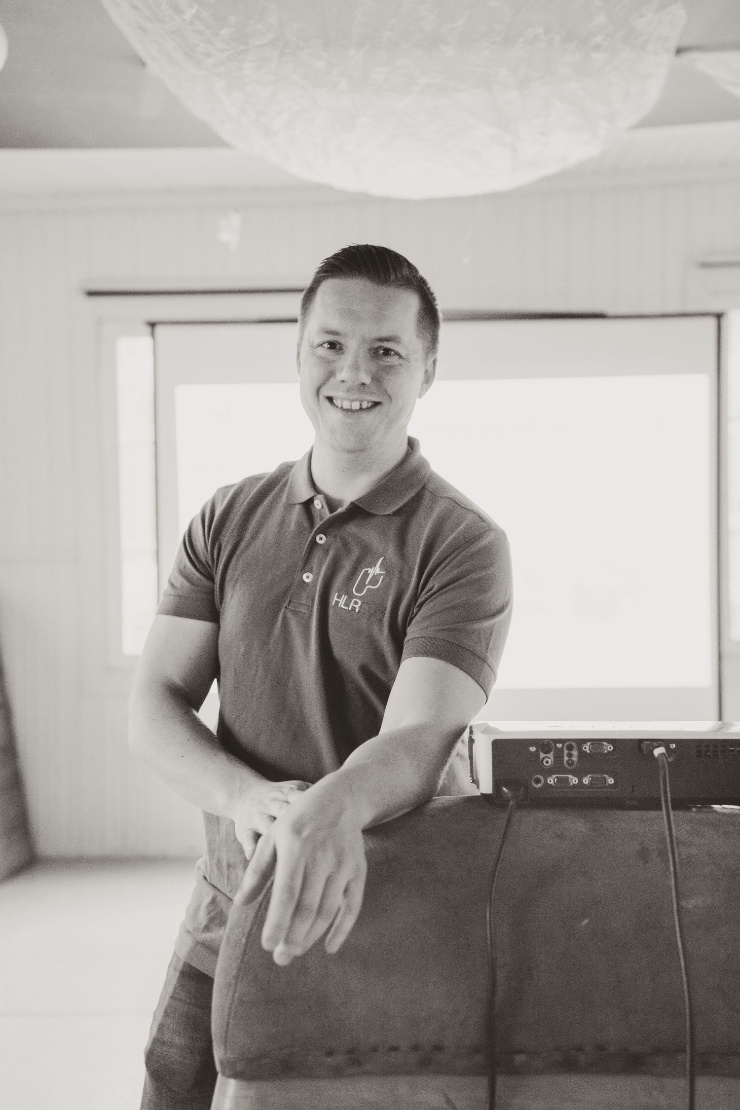 Professional in a polo shirt with logo leaning casually against furniture in bright office setting.