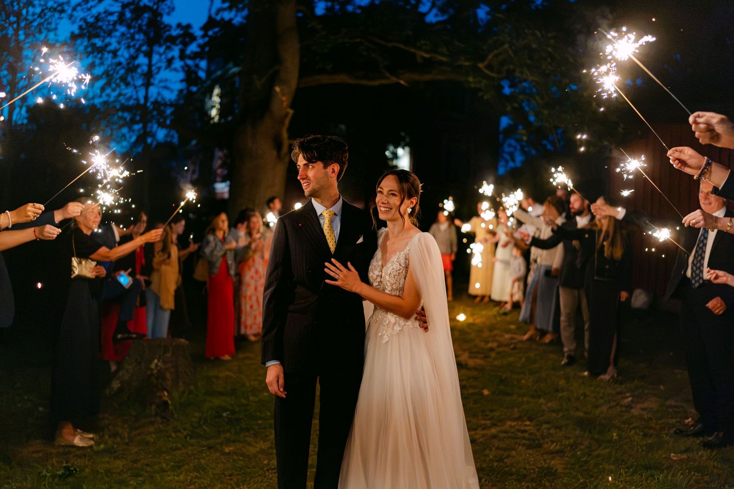 Bride and groom walk through sparkler send-off tunnel at romantic outdoor evening wedding celebration.