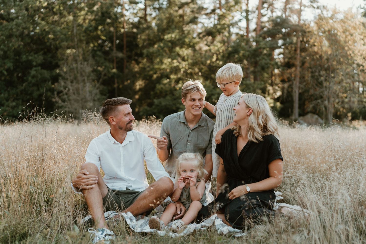 A joyful family gathering sits together in a sun-lit field with tall grass and trees in the background.