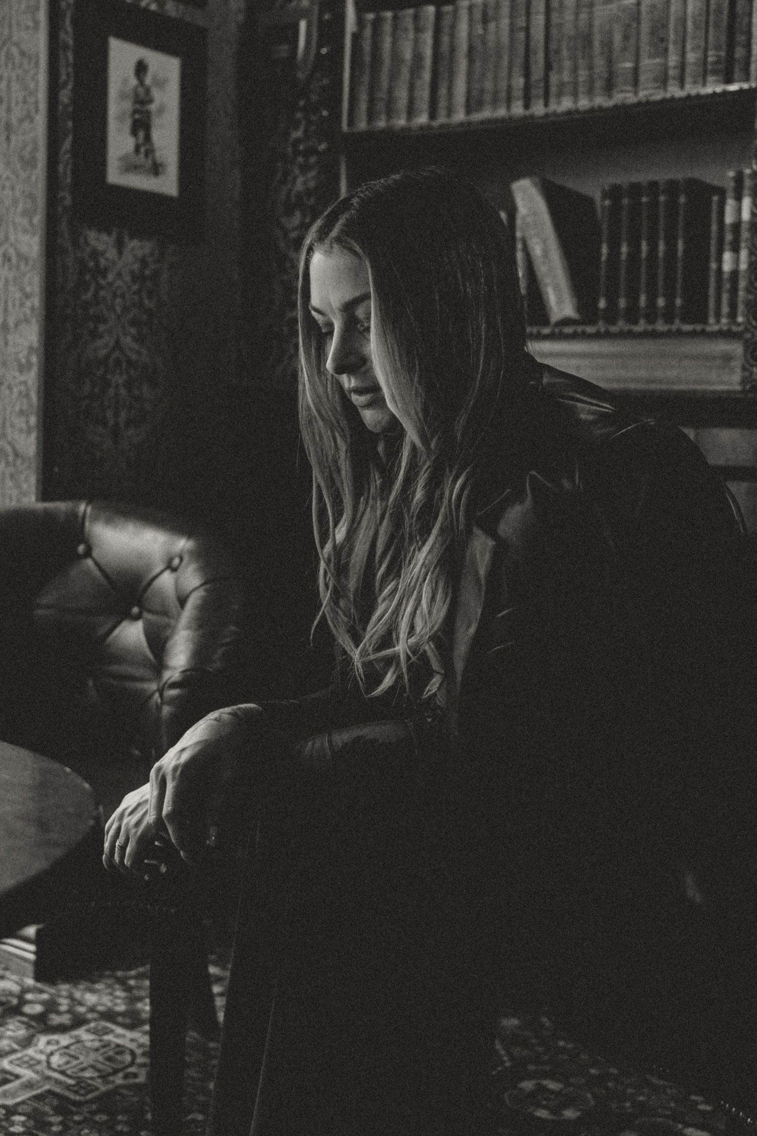 Moody black and white portrait in a library with vintage leather chair and bookshelves.