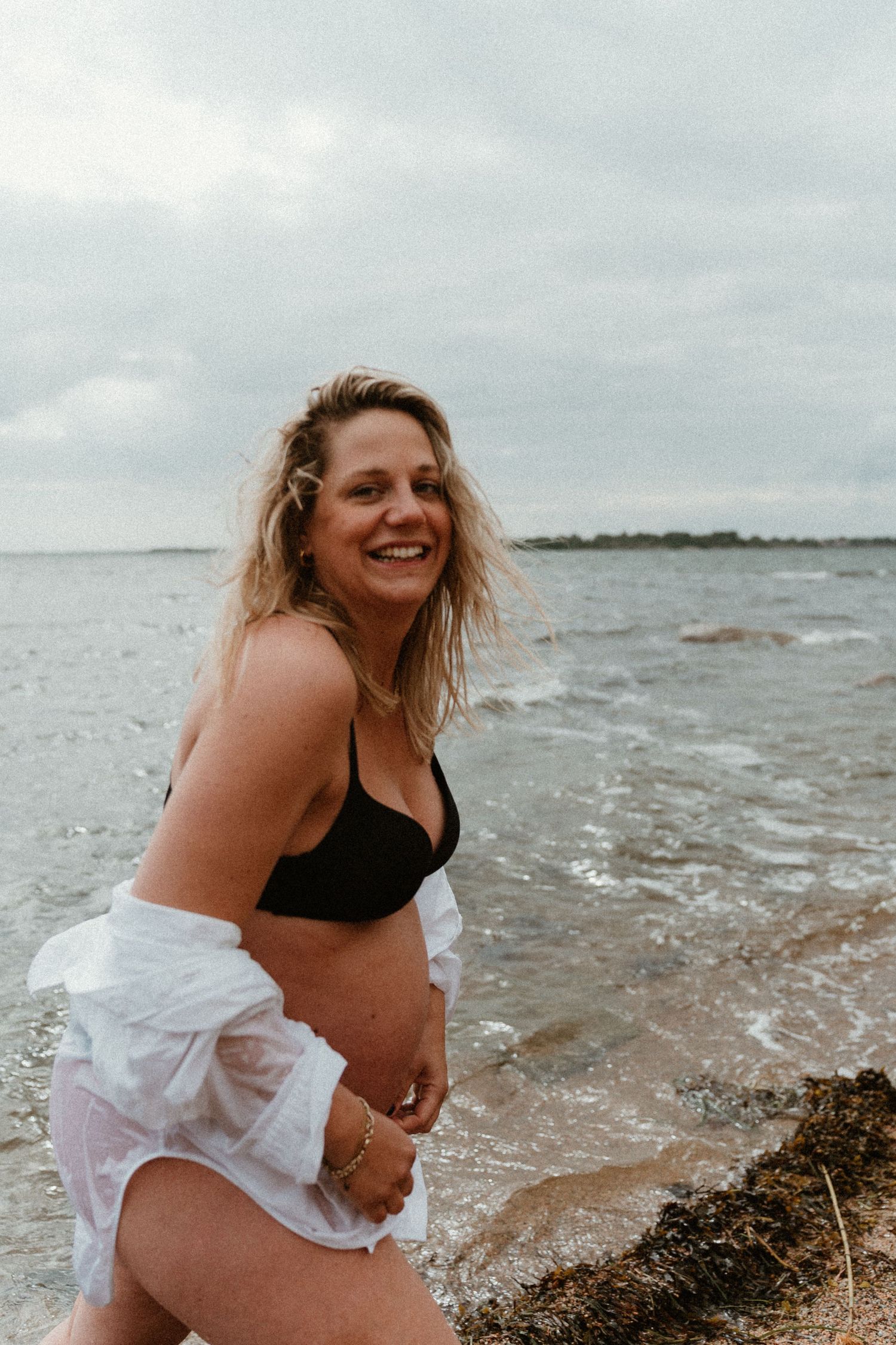 A person in a black swimsuit and white cover-up splashes and laughs joyfully at the beach on an overcast day.