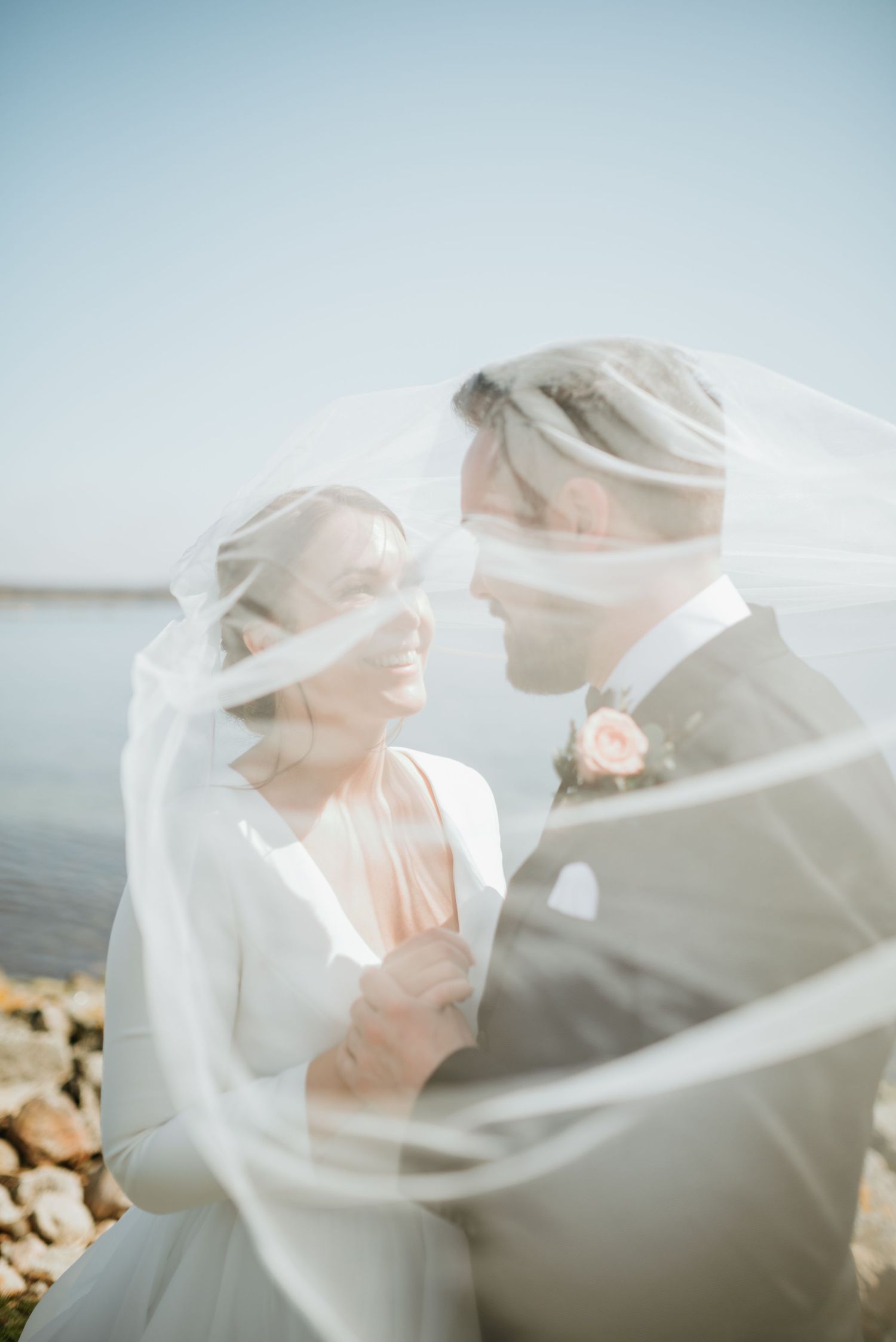 Romantic wedding portrait through flowing white veil against coastal background.