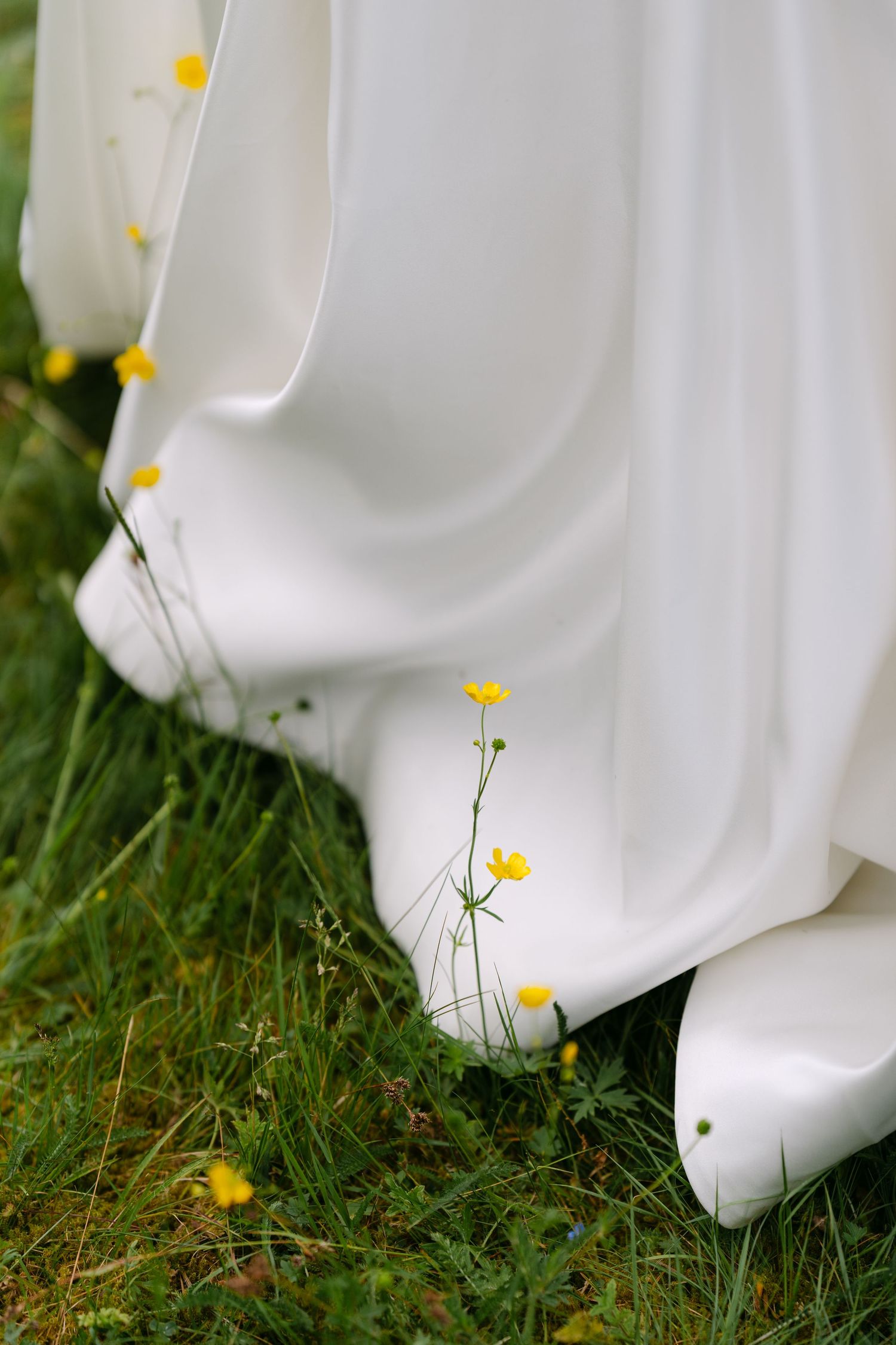 White wedding dress hem resting on green grass with yellow buttercup wildflowers in a natural outdoor setting.