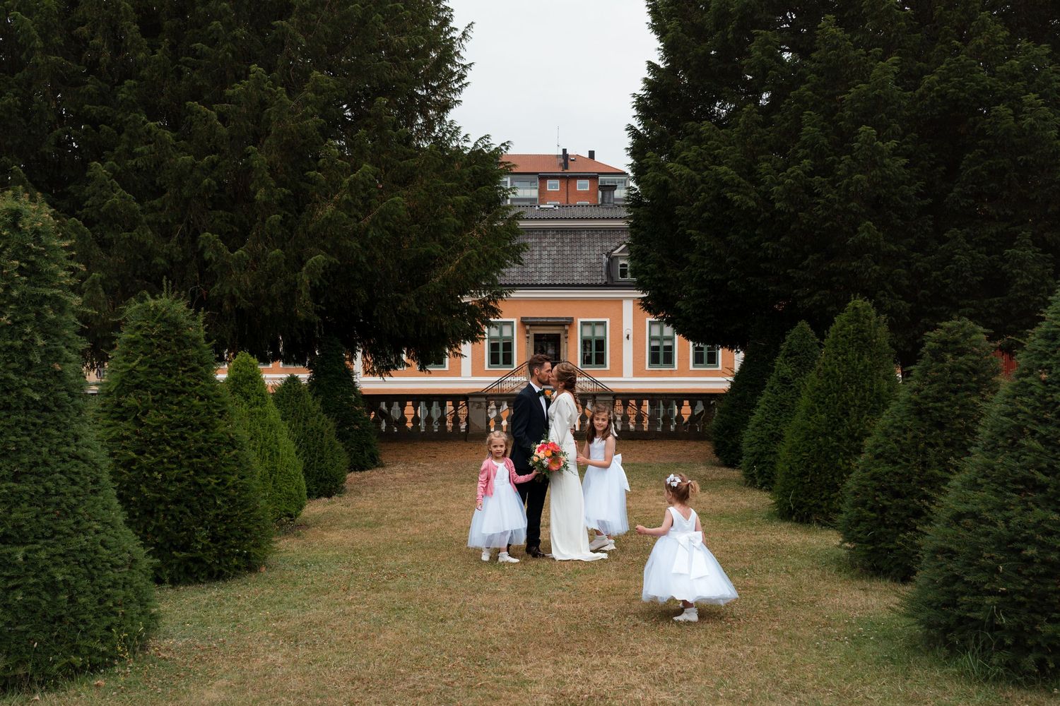 Wedding couple walks through a garden path lined with tall evergreen trees and a historic mansion in the background.