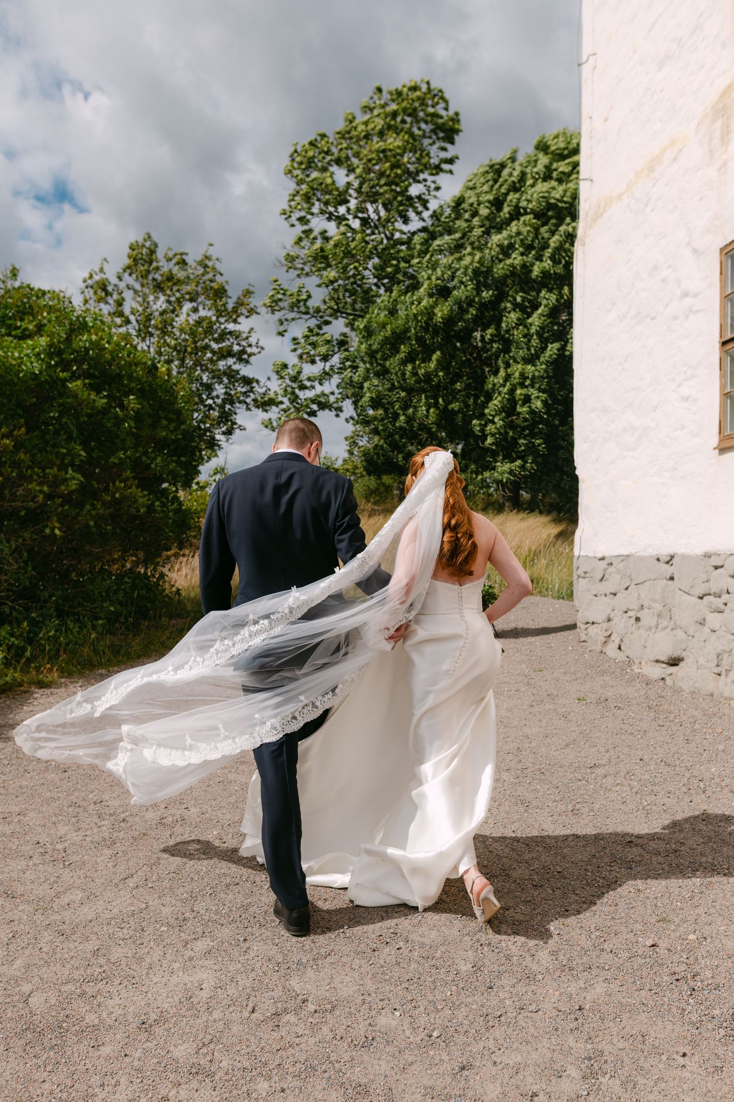 A bride and groom walk hand in hand away from camera, her lace veil billowing in the wind on a sunny wedding day.