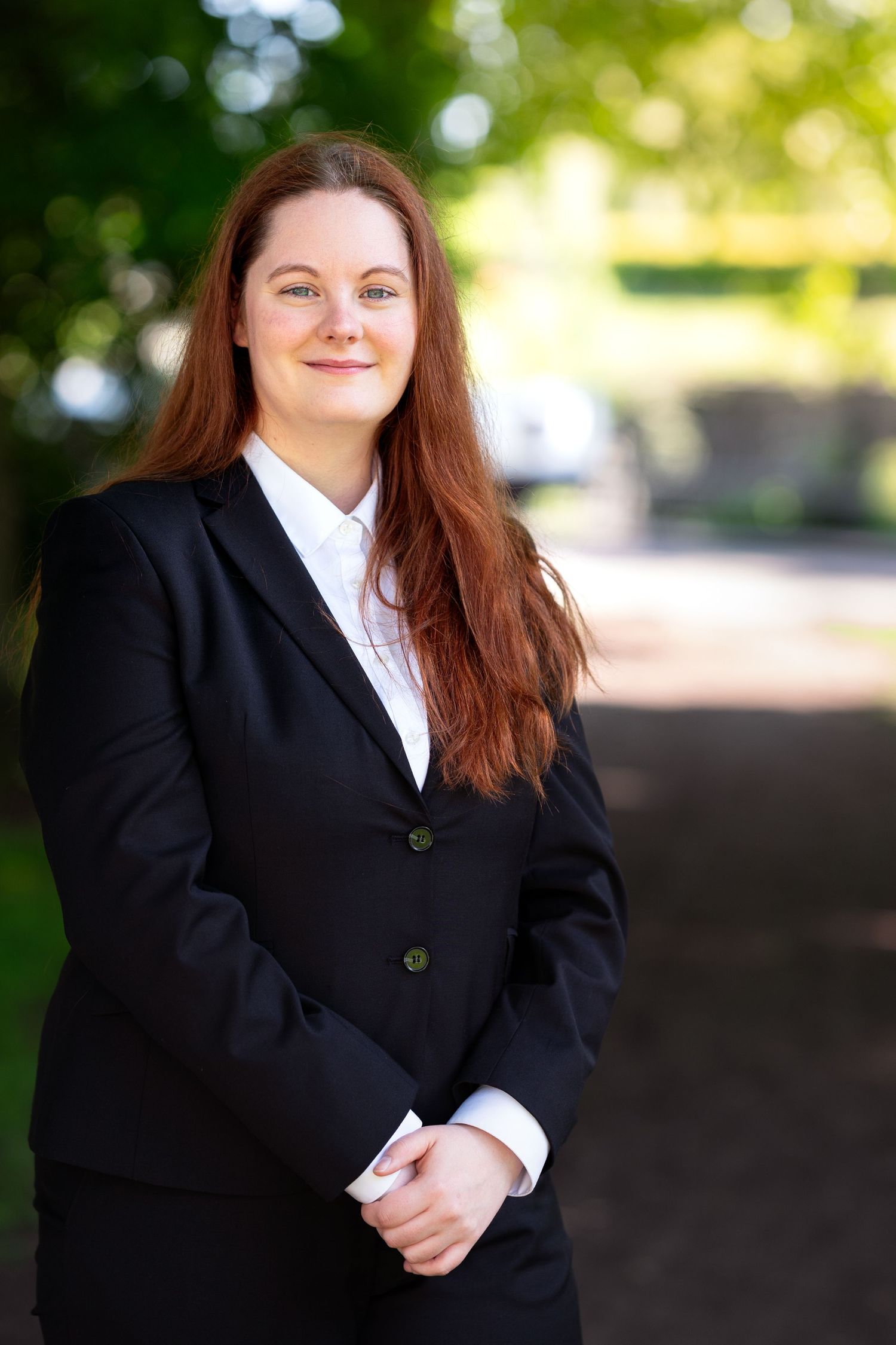 Professional portrait of someone in a black business suit and white shirt standing outdoors with a blurred green background.
