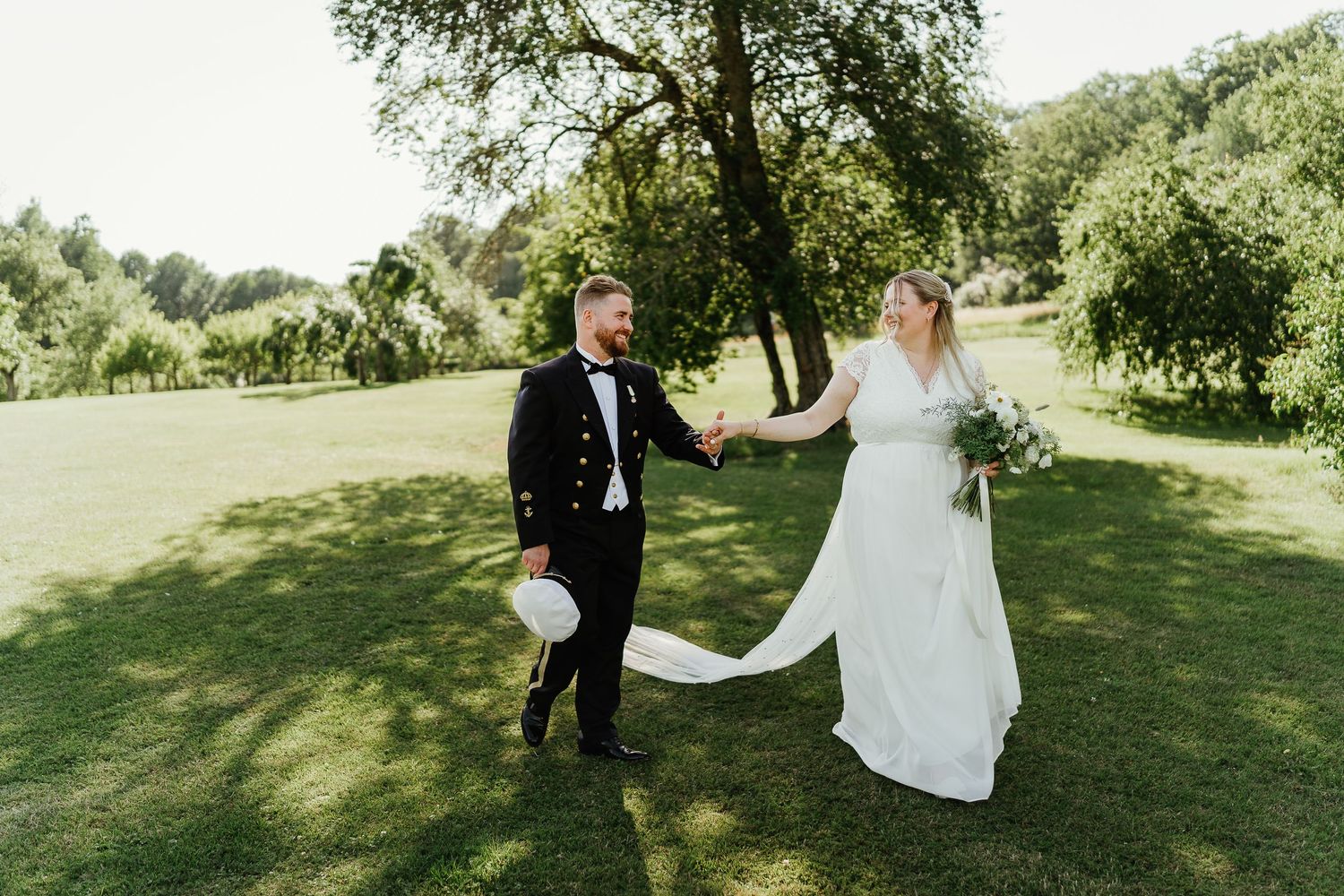 Bride and groom walk hand-in-hand on lush green lawn, bride in white gown holding bouquet, groom in black suit.