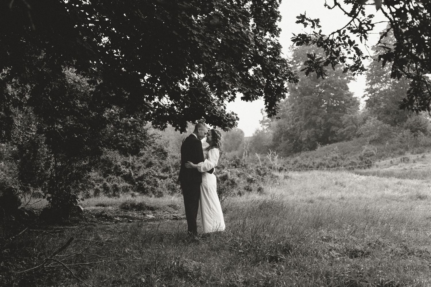 A romantic black and white wedding portrait of a couple embracing under a large tree in a meadow.