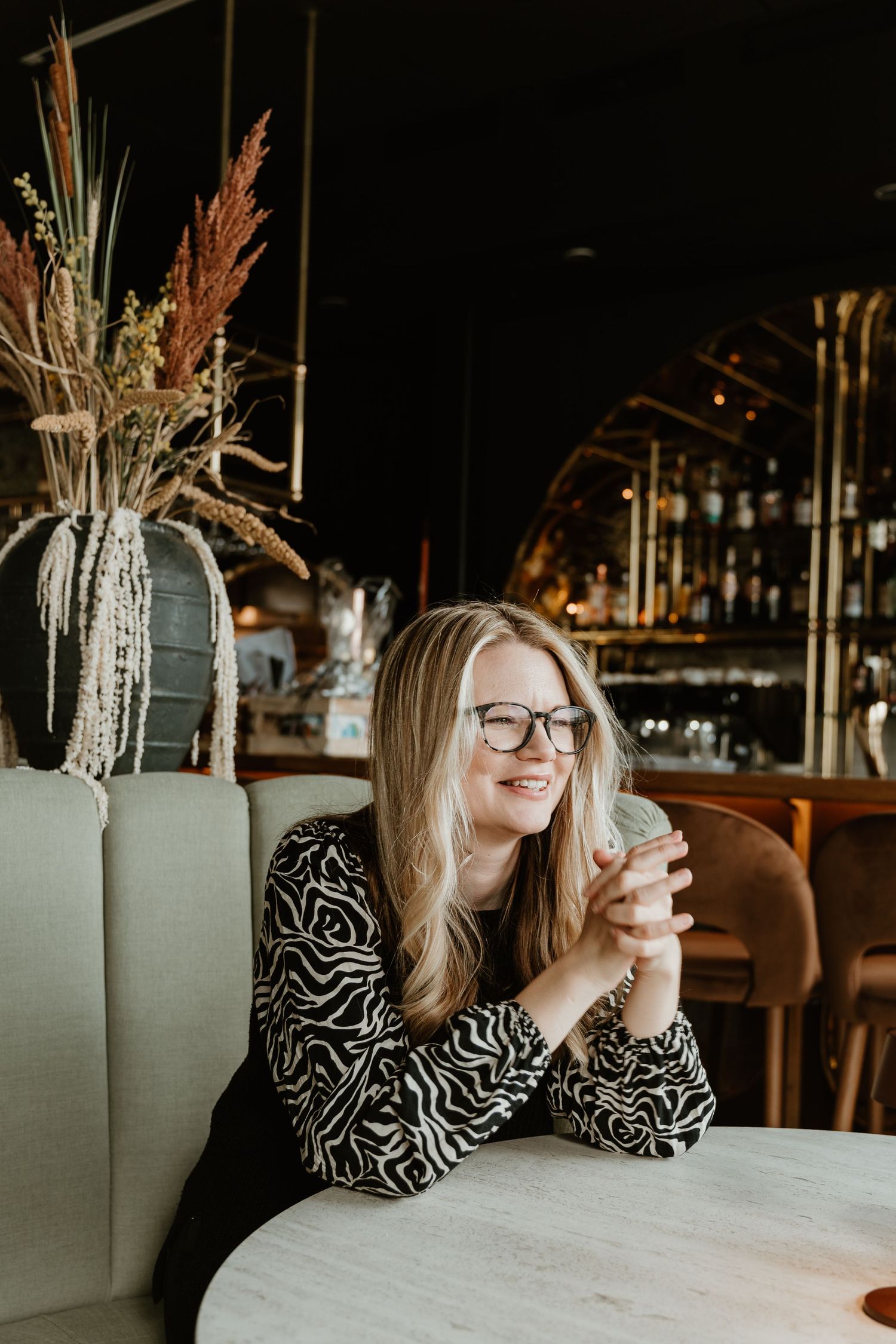 A candid portrait of someone wearing glasses and a patterned top relaxing at a cafe with decorative dried flowers.