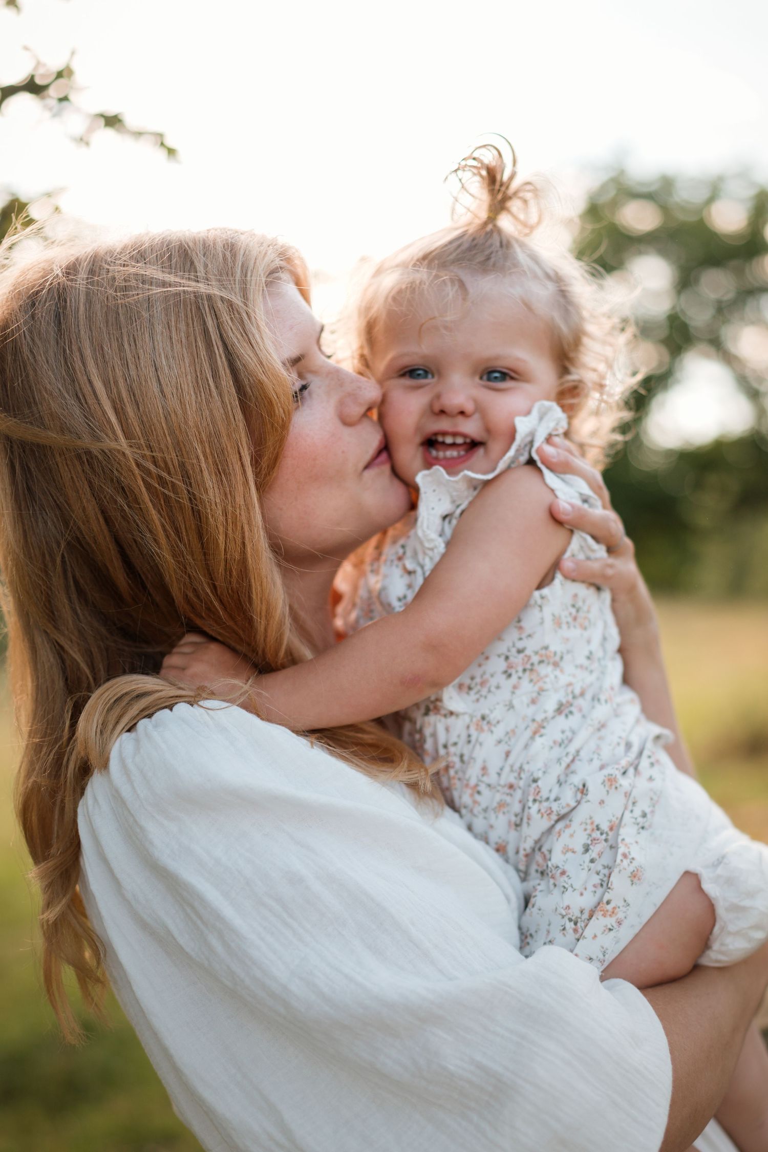 A mother tenderly kisses her smiling toddler daughter outdoors in golden sunlight, both wearing white outfits.