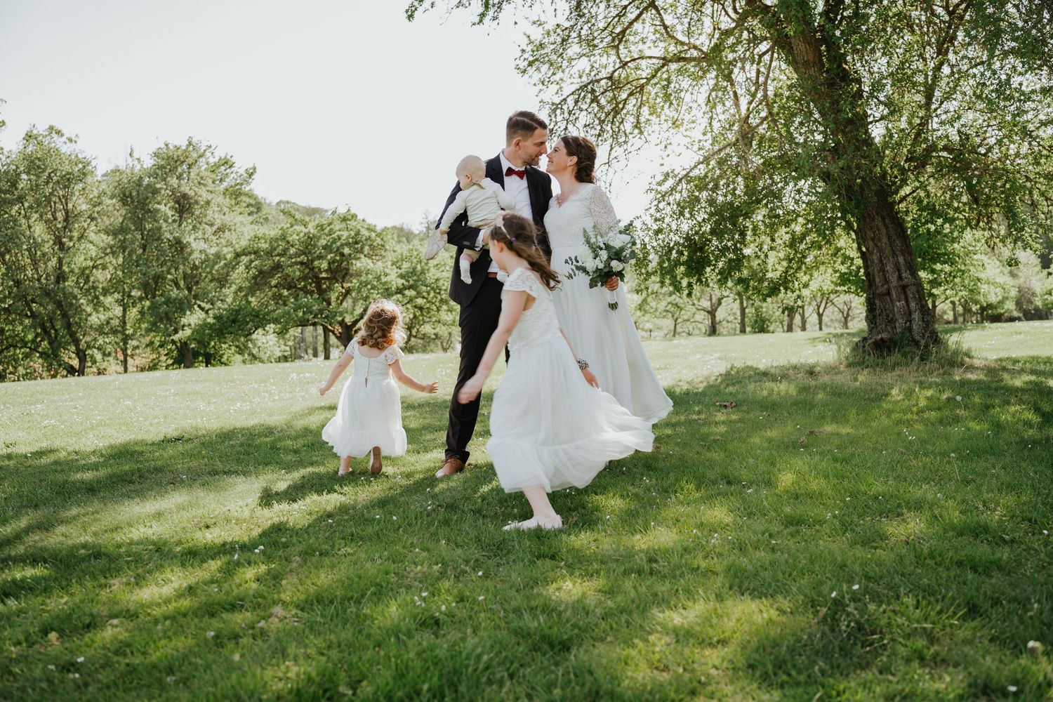 A joyful wedding moment captures a couple dancing on green grass beneath a tree while a small child joins in white dress.