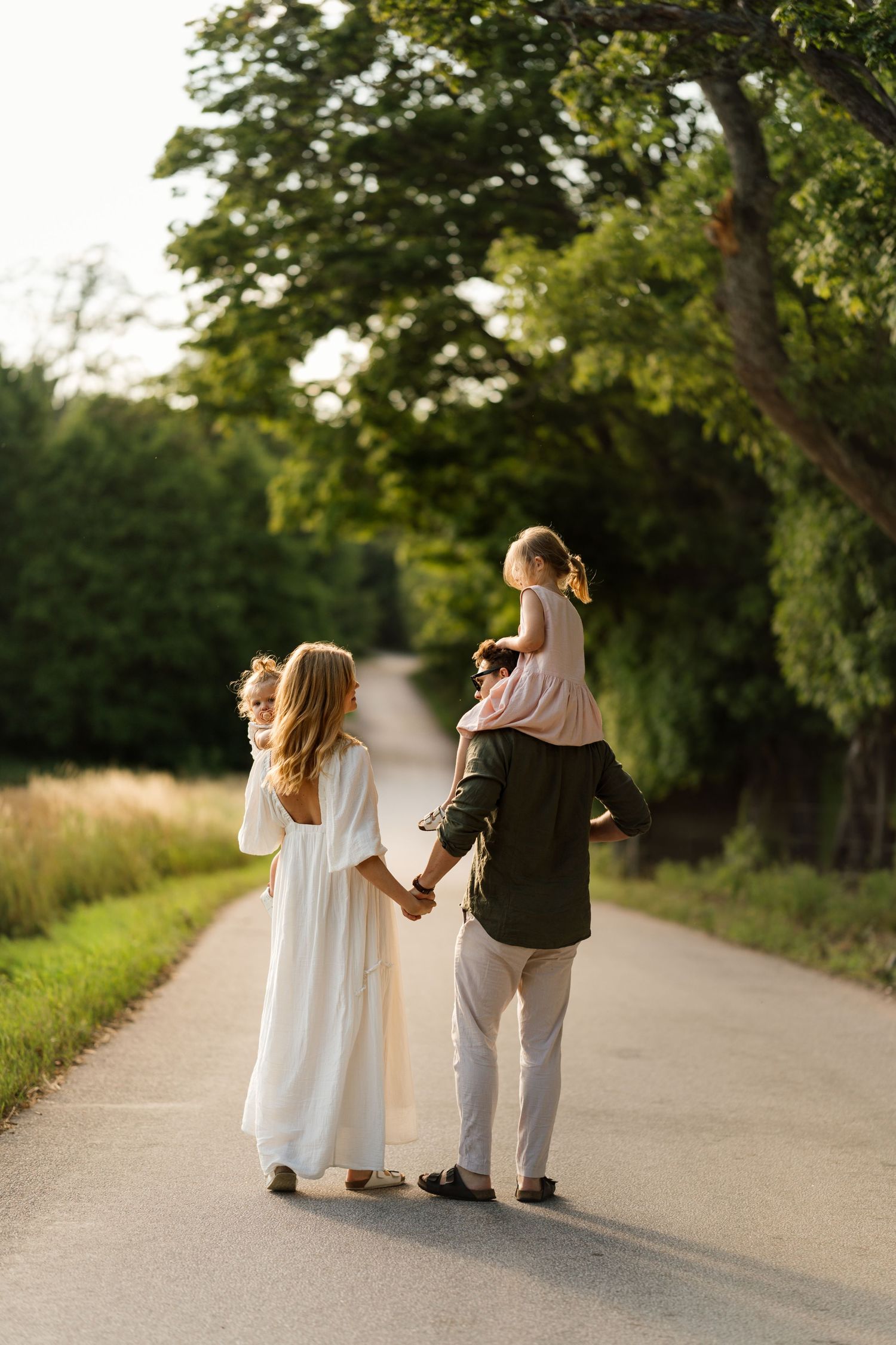 Family walking hand-in-hand down a sunlit road, with a child on dad's shoulders, surrounded by lush green trees.