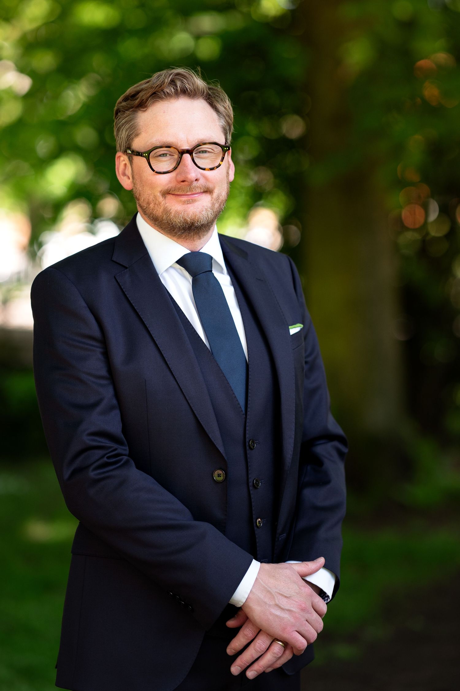 Professional portrait series of someone in a navy suit and glasses against a blurred green outdoor background.