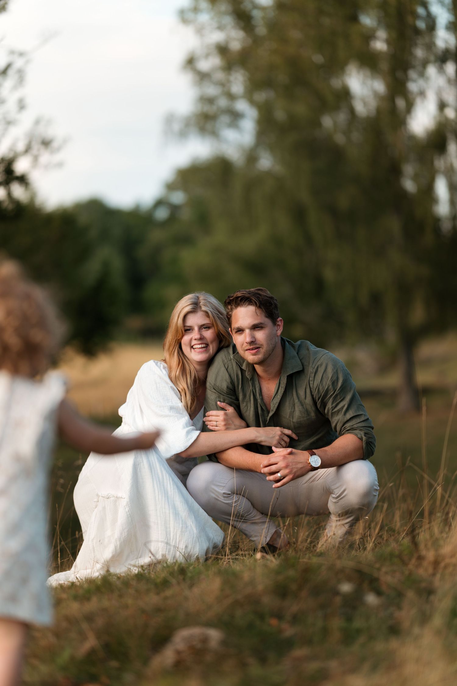 A smiling couple crouches in a grassy field while a young child approaches them on a warm summer day.