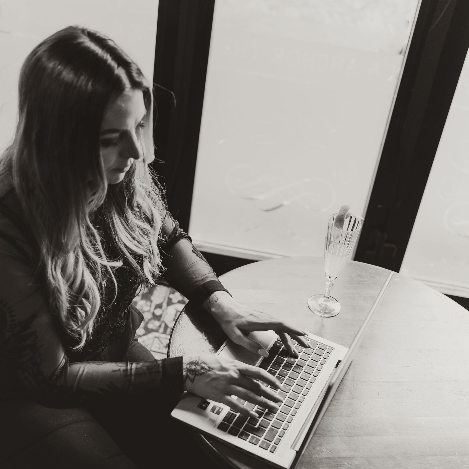 Person typing on laptop with champagne glass on table in monochrome setting.