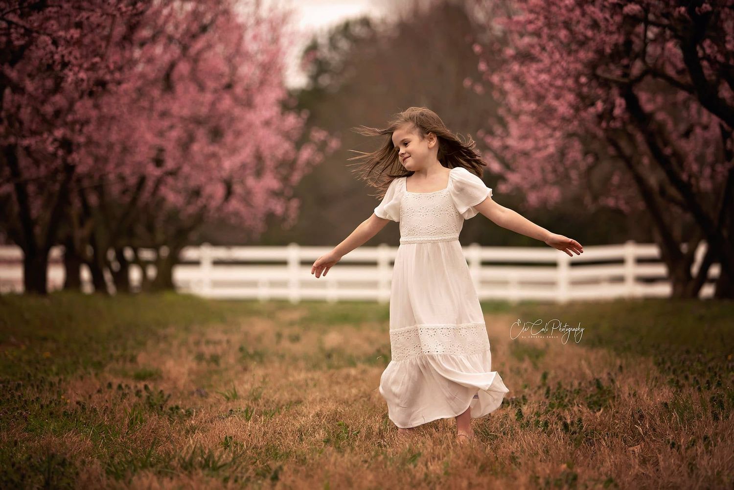 A young girl twirls freely in a field lined with blooming pink trees, her white dress flowing softly around her.