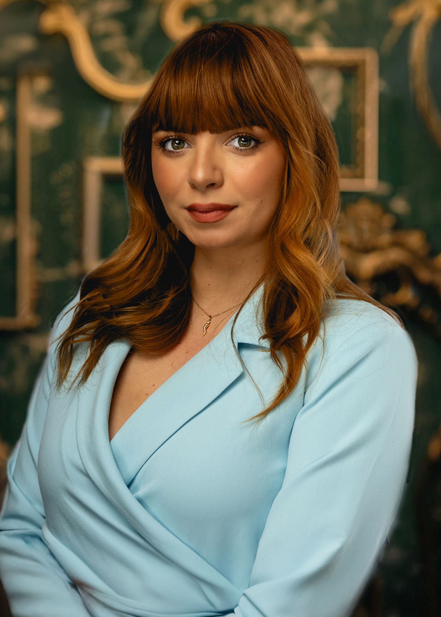 Professional portrait of someone wearing a light blue blazer against an ornate patterned background in warm lighting.