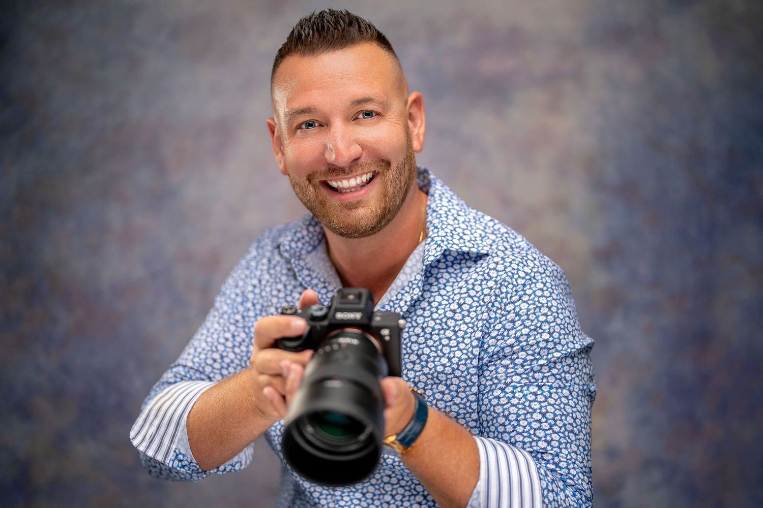 Professional photographer wearing blue patterned shirt holding DSLR camera against studio backdrop.