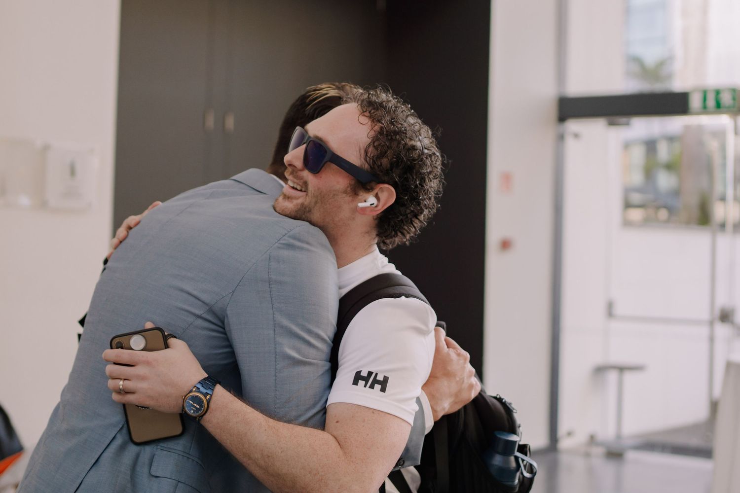 Two men sharing a warm hug indoors, one wearing sunglasses and a Helly Hansen shirt.