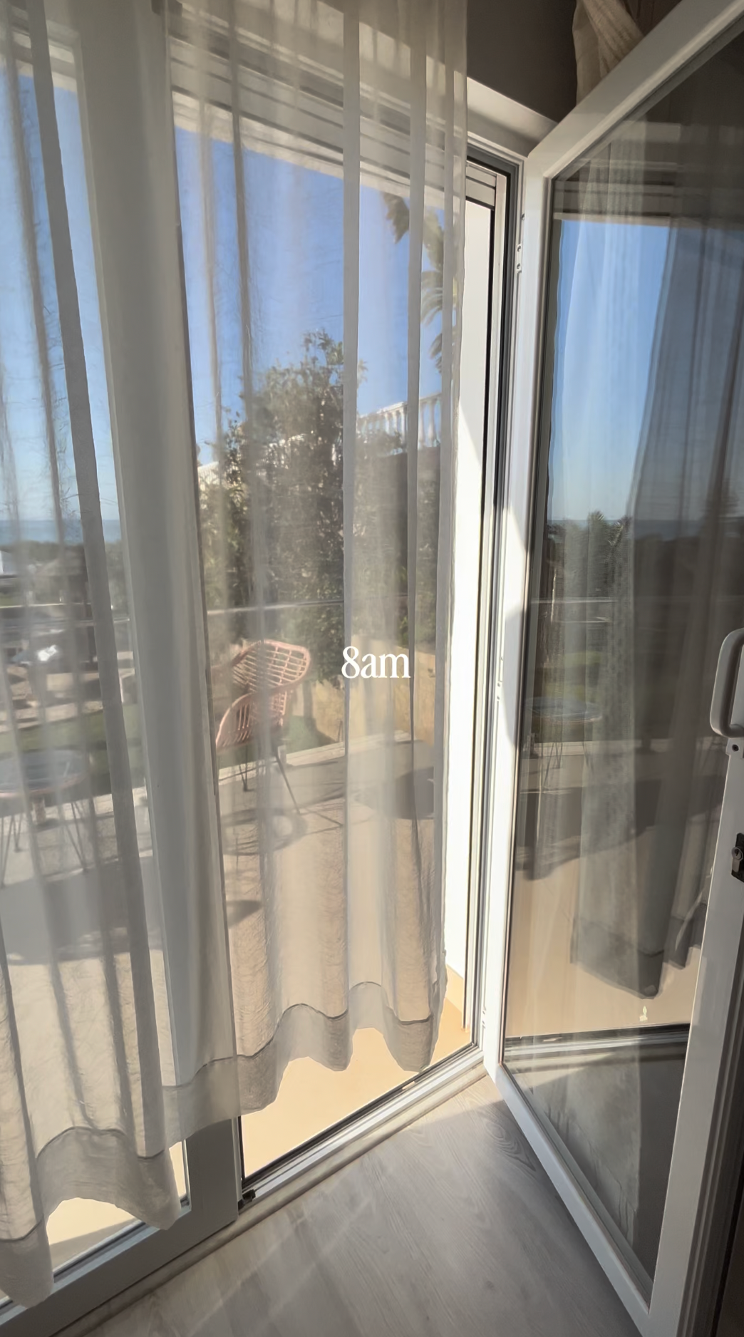 Sheer white curtains frame a sliding glass door opening to a sunny terrace with trees and outdoor furniture.