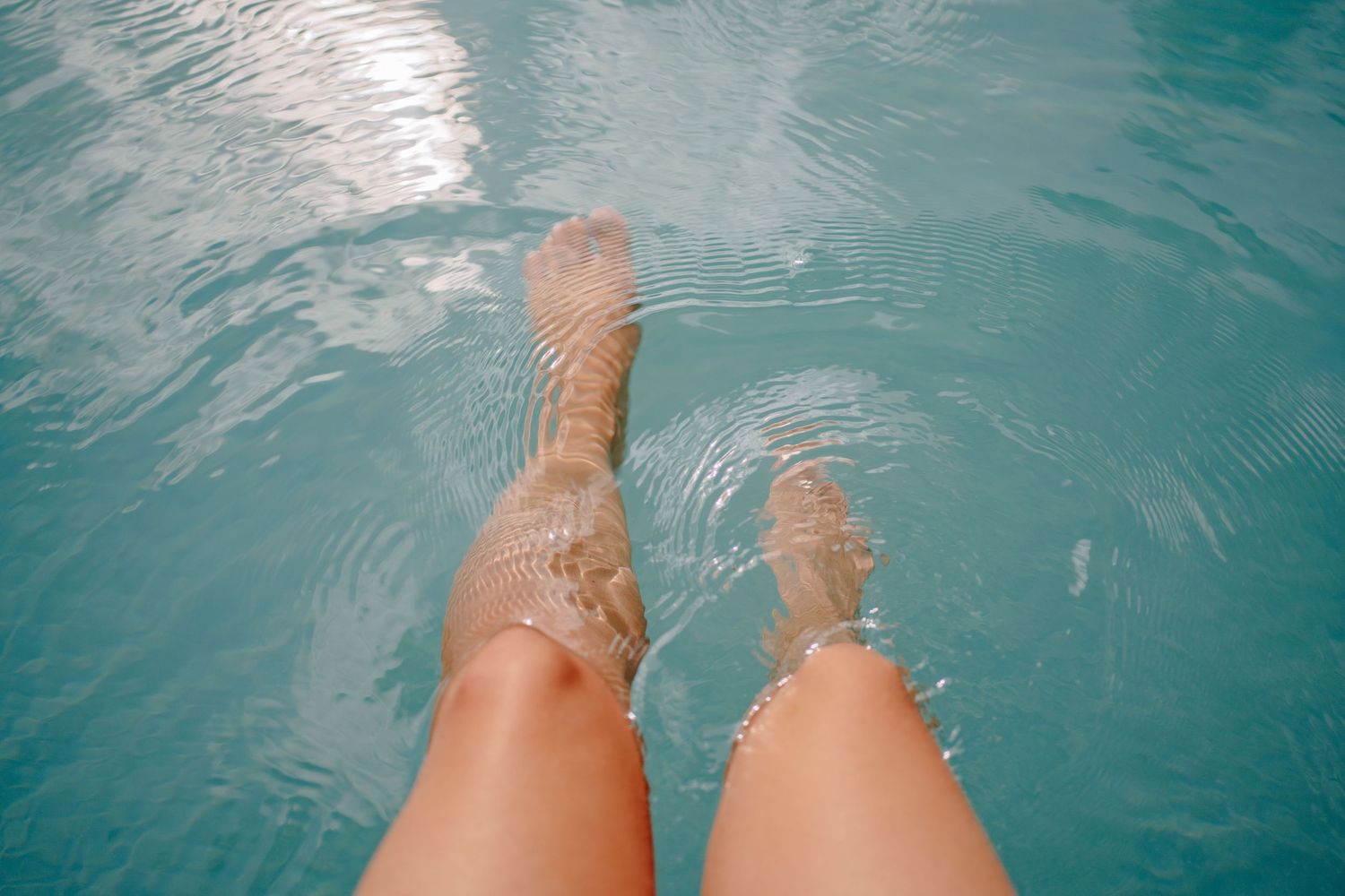 Person's bare legs and feet dangling in clear turquoise pool water, creating gentle ripples and sunlight reflections on the surface.