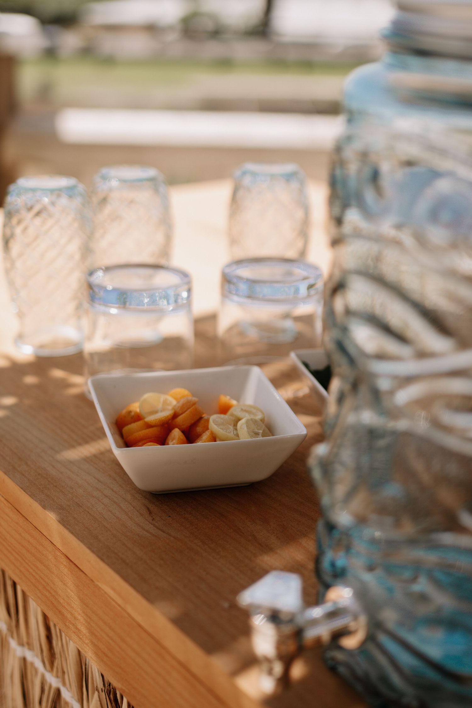 Rustic wooden bar with mason jar glasses, a glass drink dispenser, and a bowl of lemon slices in warm sunlight.
