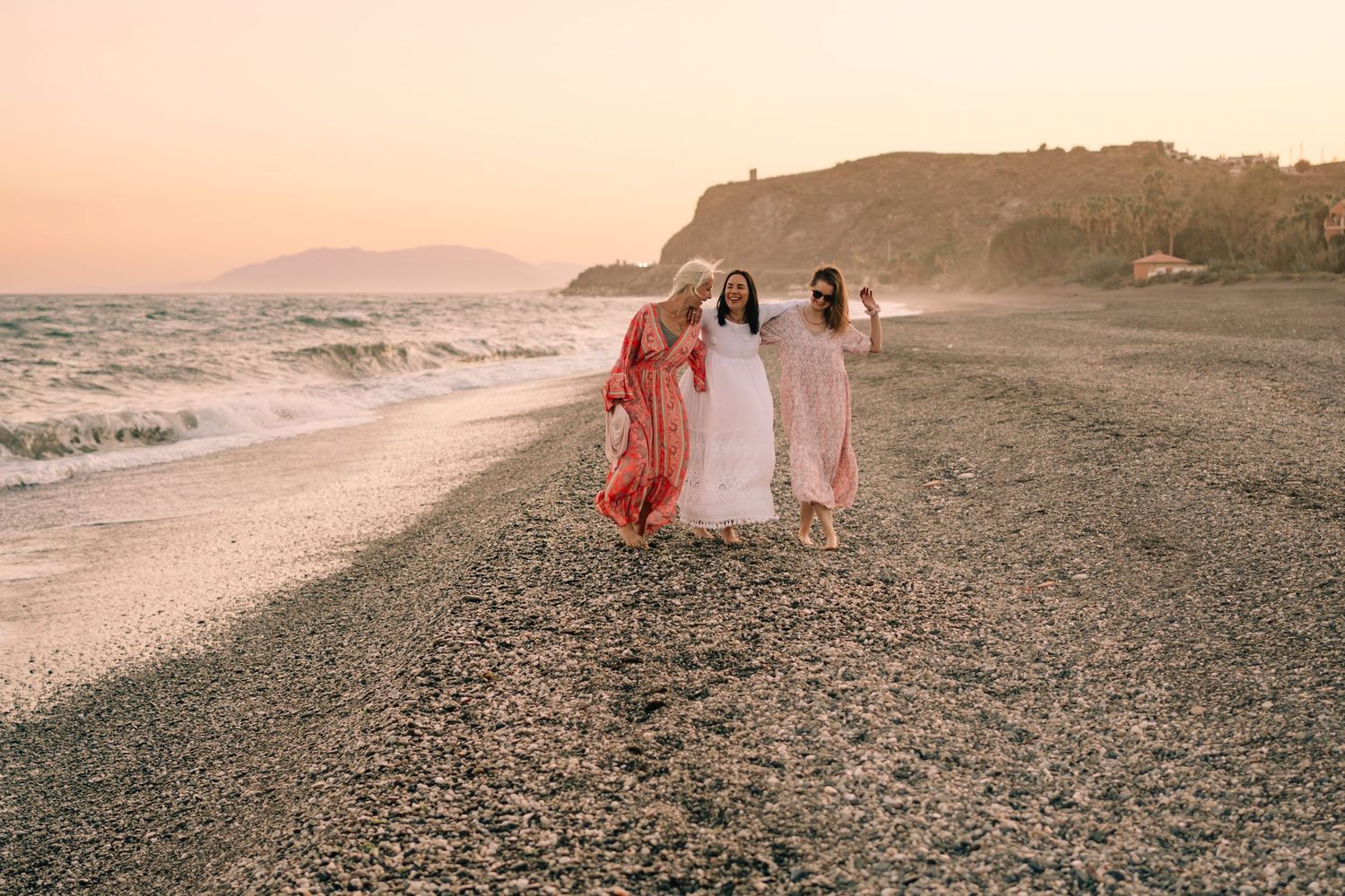 Three women in flowing dresses walking on a rocky beach at sunset with mountains in the background.
