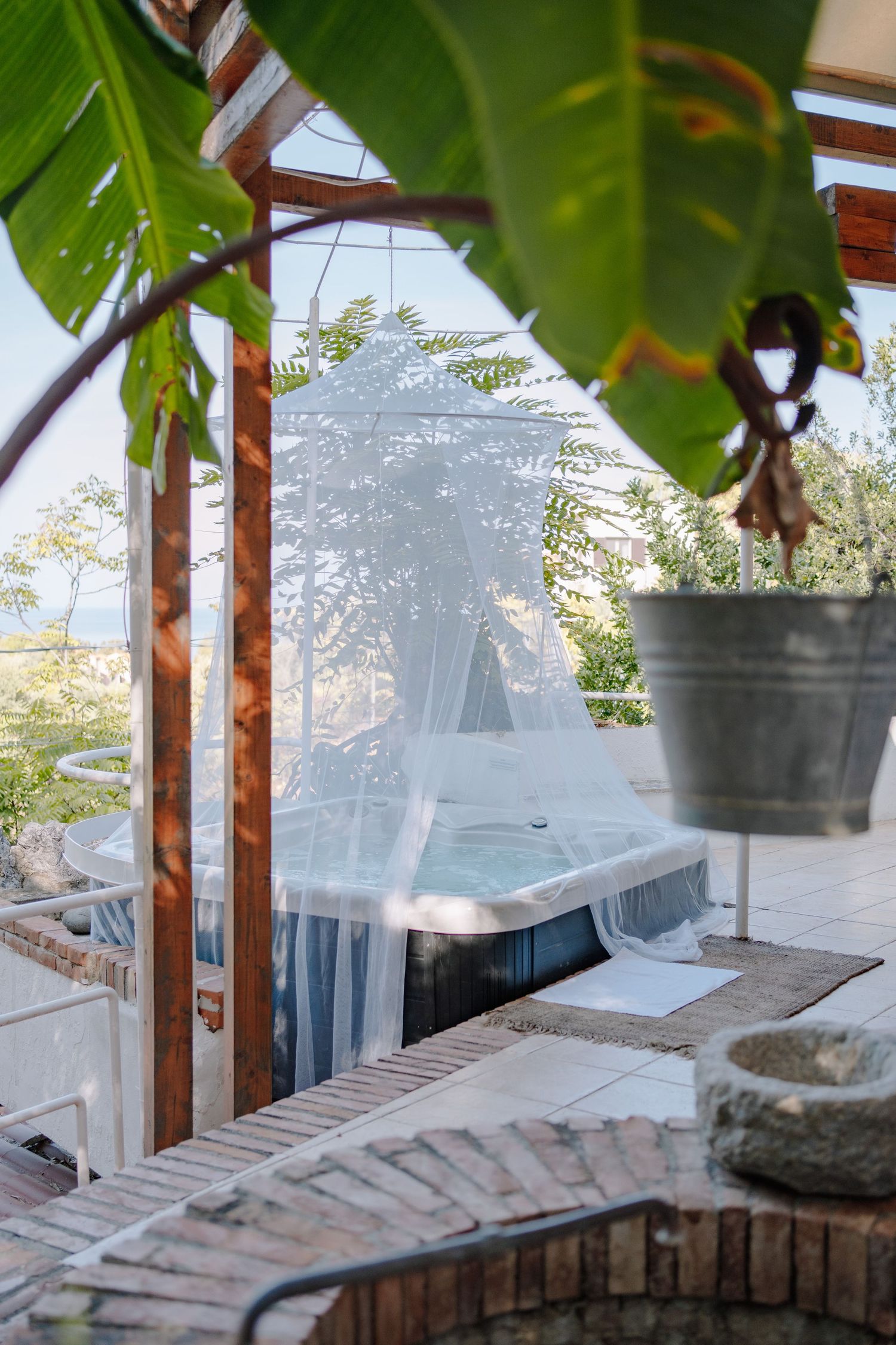 Outdoor hot tub covered by a white mosquito net under a wooden pergola on a sunny brick terrace with tropical leaves.