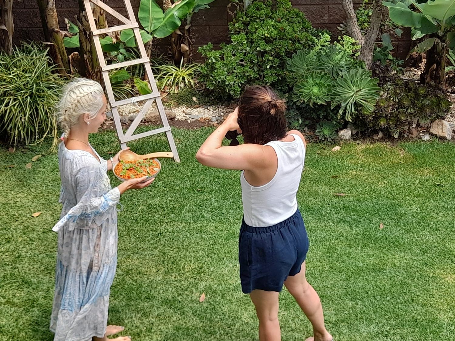 Two girls in a sunny backyard garden, one holding a bowl near a wooden ladder surrounded by tropical plants.