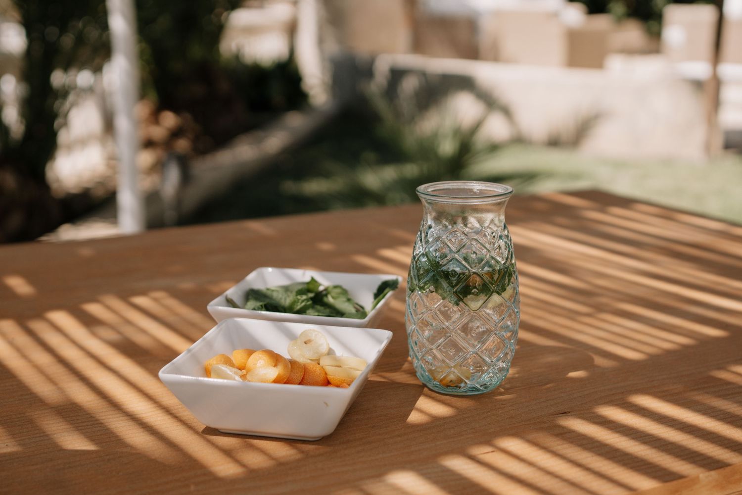 Glass pitcher with mint water and white bowls of fresh fruit and greens on a sunny wooden outdoor table.