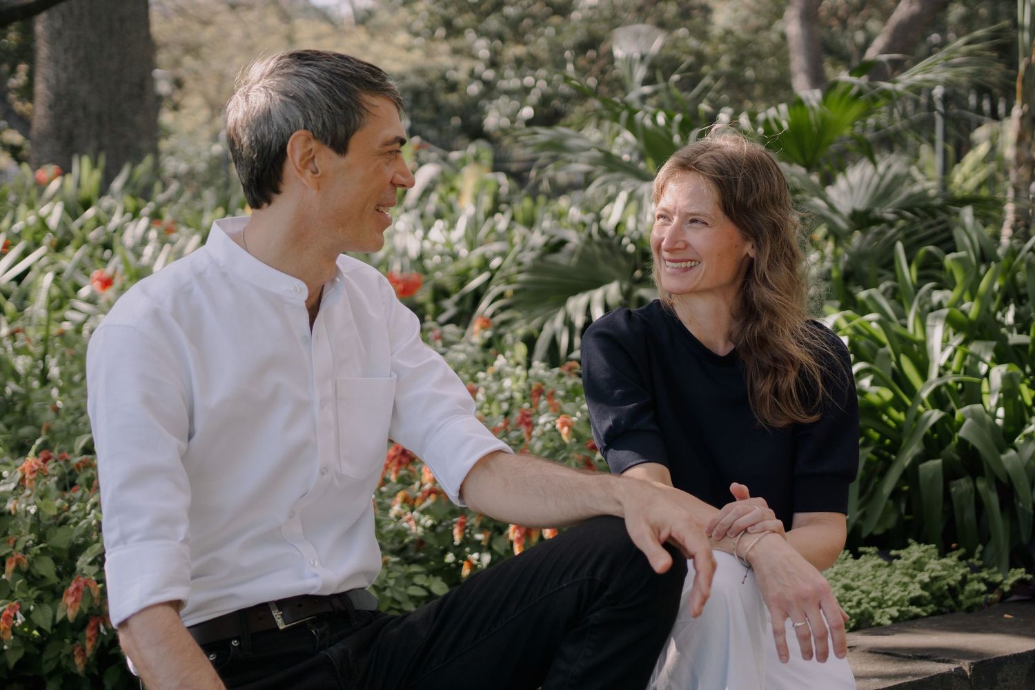 A man and woman sit together smiling in a lush garden, wearing casual attire in a warm, natural setting.