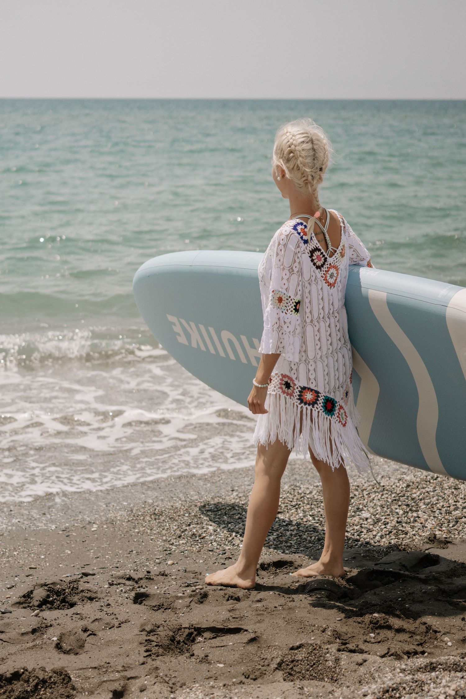 Blonde woman in white crochet cover-up carrying a blue paddleboard on a rocky beach with turquoise ocean waves.