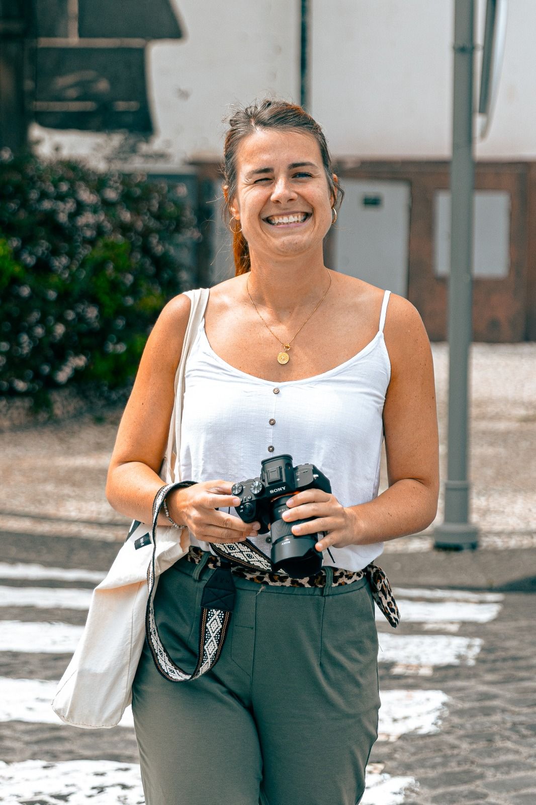Smiling woman holding a camera outdoors, wearing a white tank top and green pants with a bag over her shoulder.
