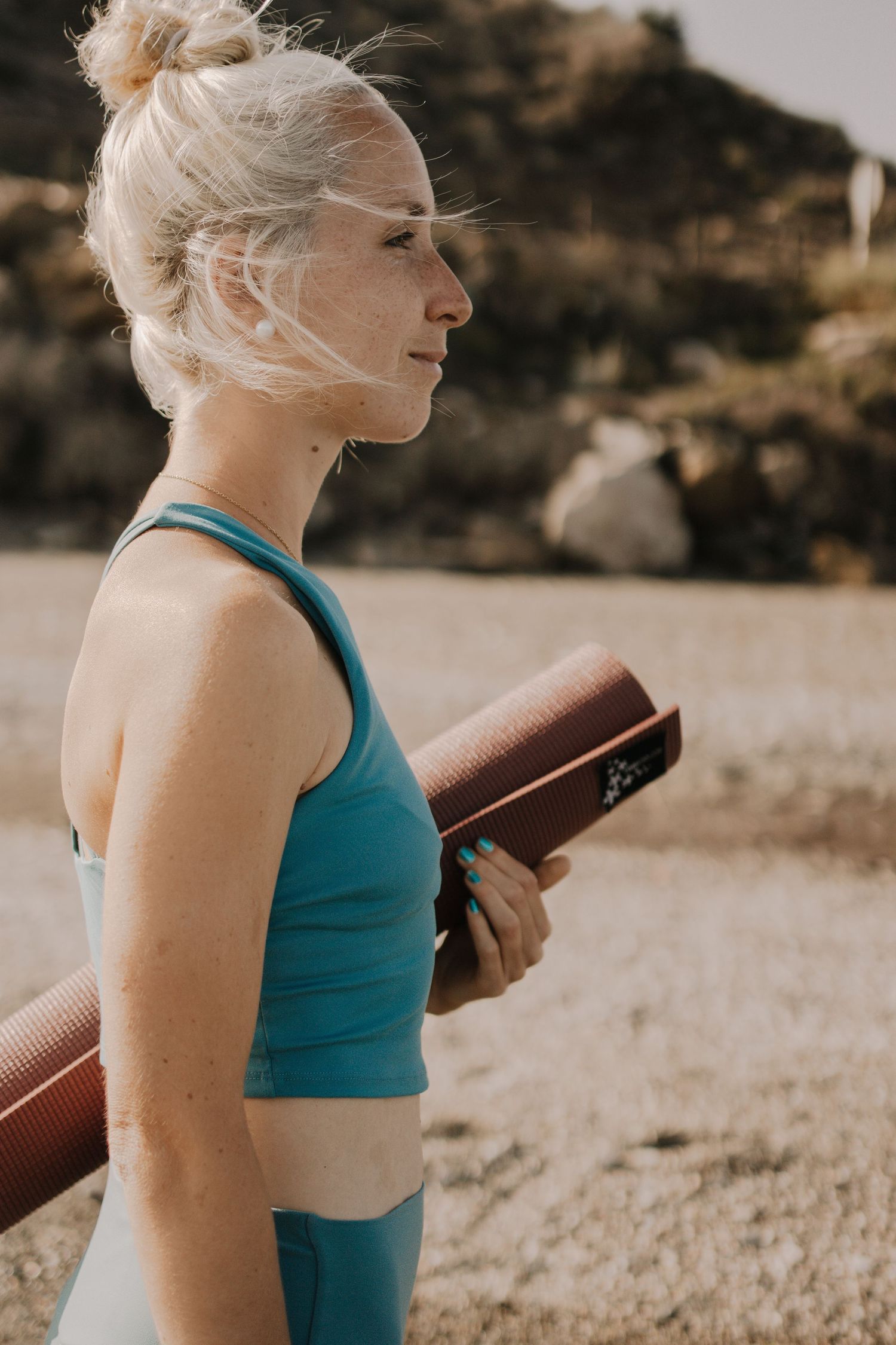 A blonde woman in teal activewear holds a rolled yoga mat outdoors in a sunny, arid landscape.