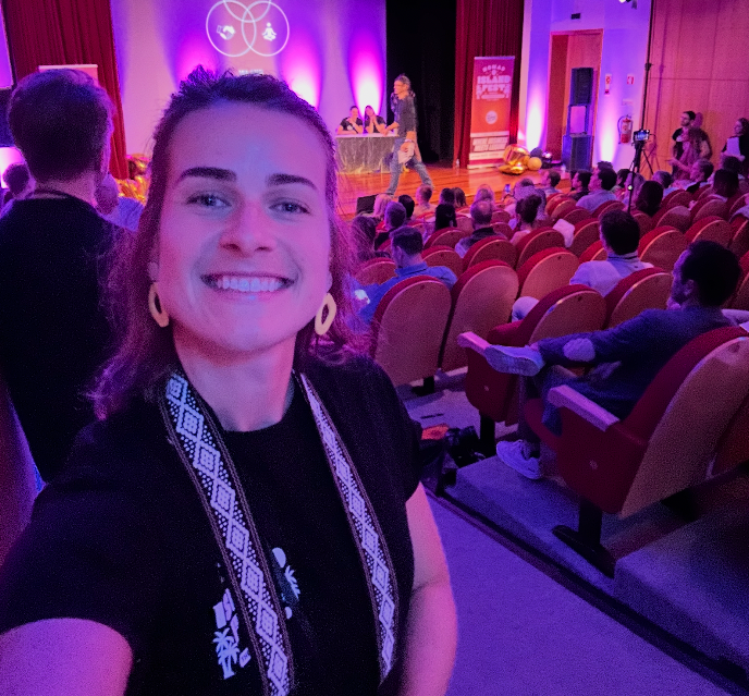 Woman smiling for selfie at conference event with audience and stage presentation visible in background.