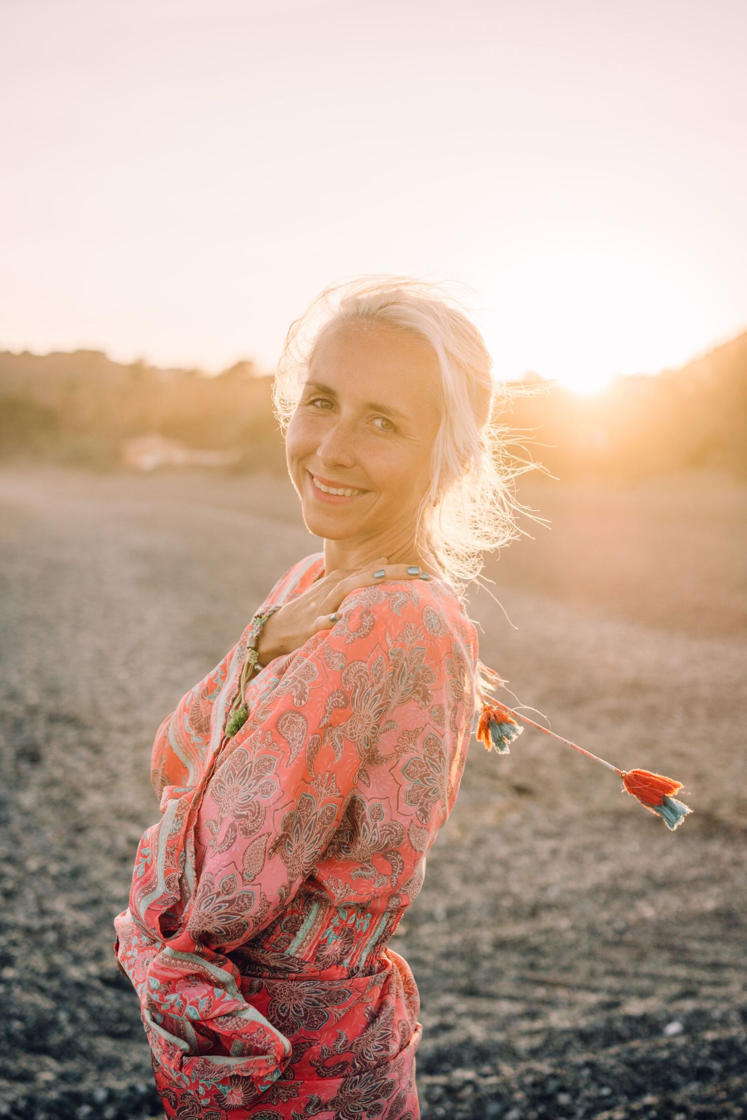 Smiling blonde woman in pink paisley dress standing on beach during golden sunset.