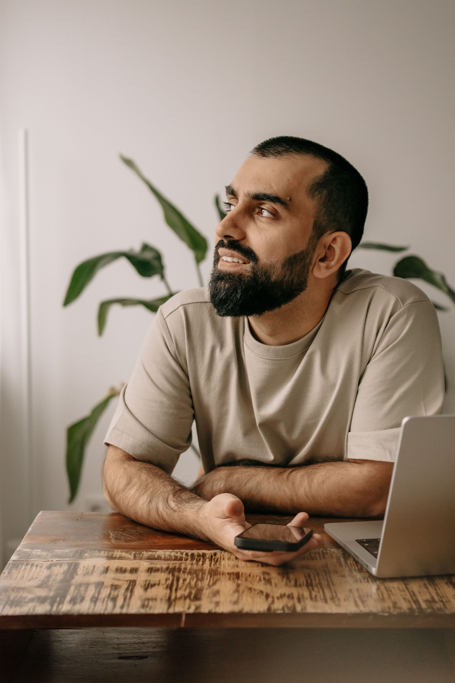 Smiling bearded man sitting at wooden desk with smartphone and laptop, looking away thoughtfully, green plant in background.