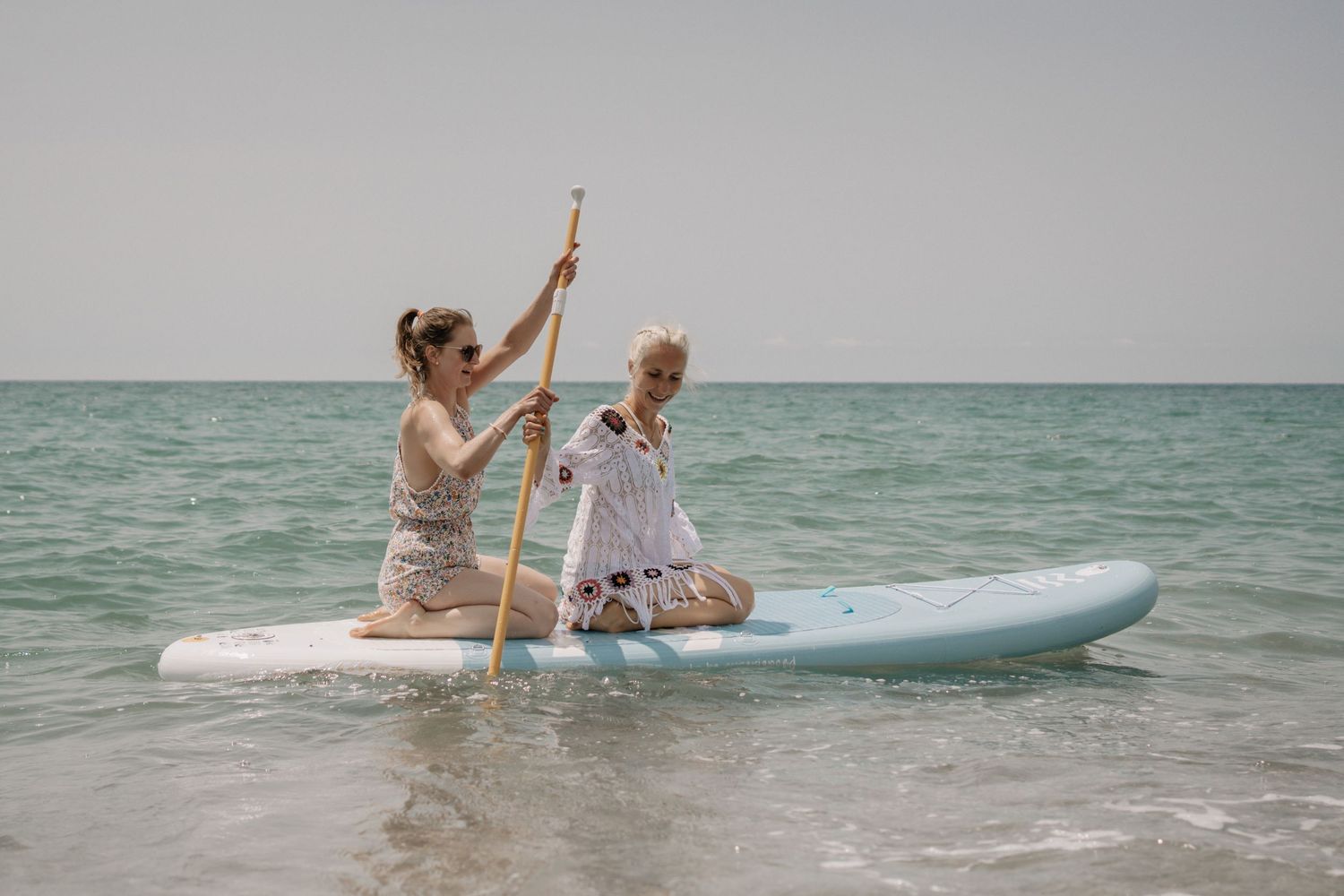 Two women paddleboarding on calm turquoise ocean waters, one holding a wooden paddle on a light blue SUP board.