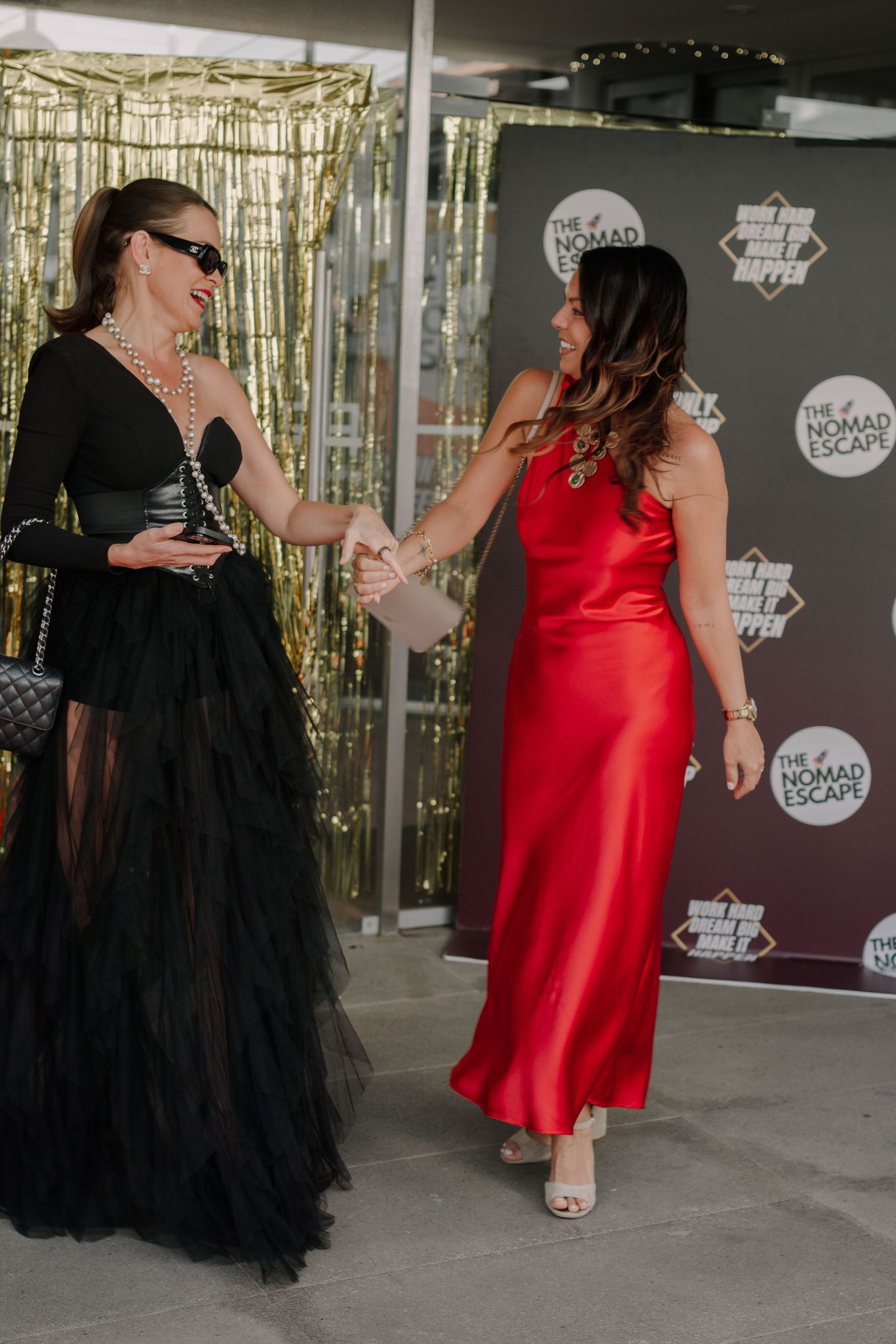 Two elegantly dressed women shaking hands at The Nomad Escape event, one in black gown and one in red satin dress.
