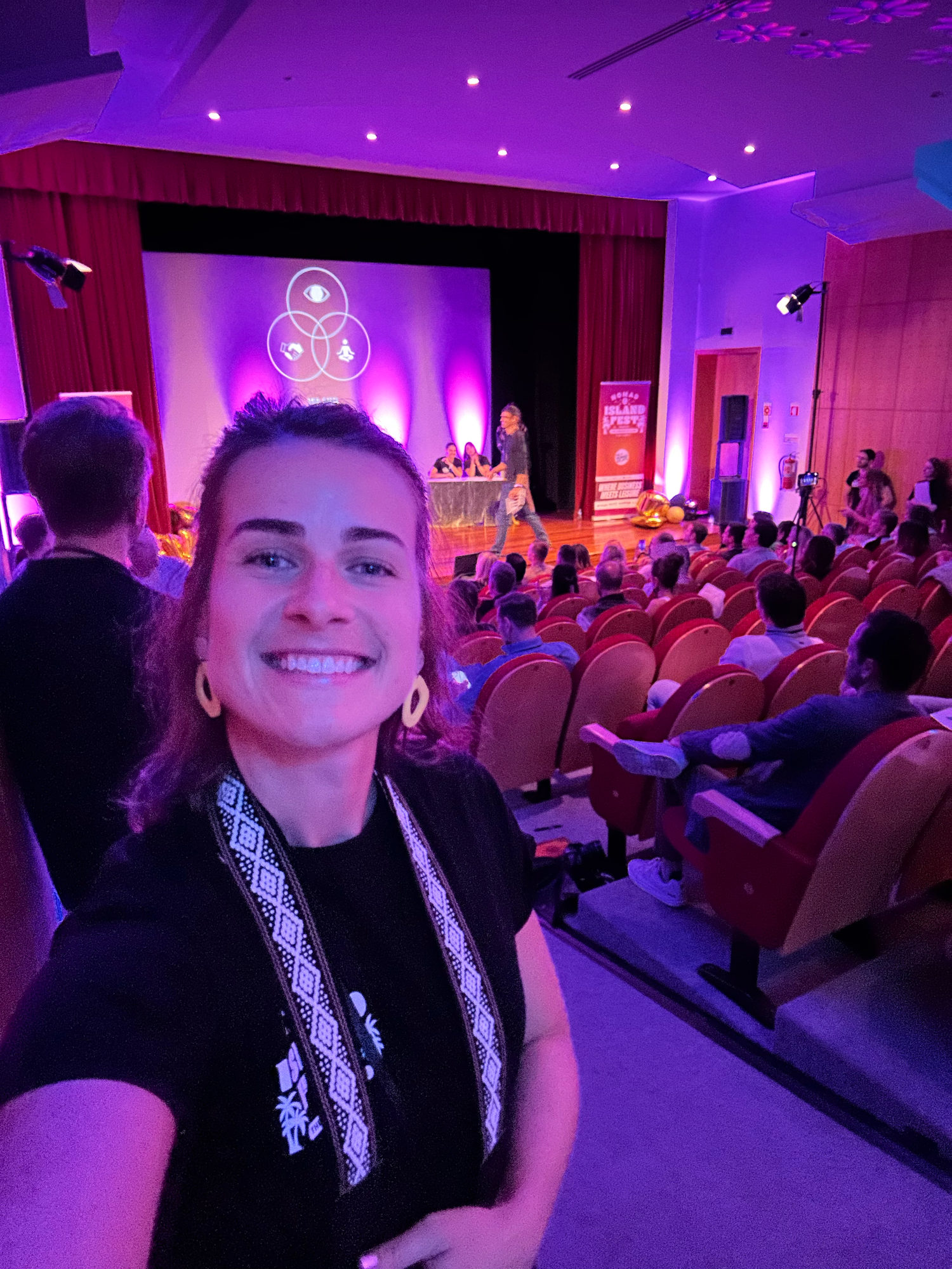Woman taking selfie at wellness conference with speaker on stage and purple lit auditorium.
