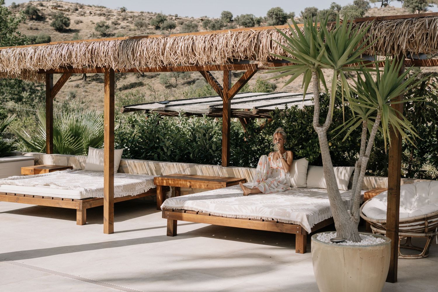 Woman relaxing on luxury outdoor daybed under thatched pergola at Mediterranean resort with lush greenery.