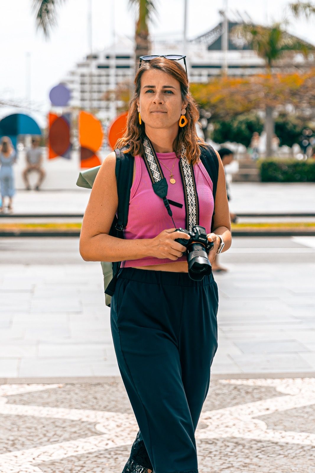 Woman in pink top holding camera with decorative patterned plaza and colorful art installations in background.