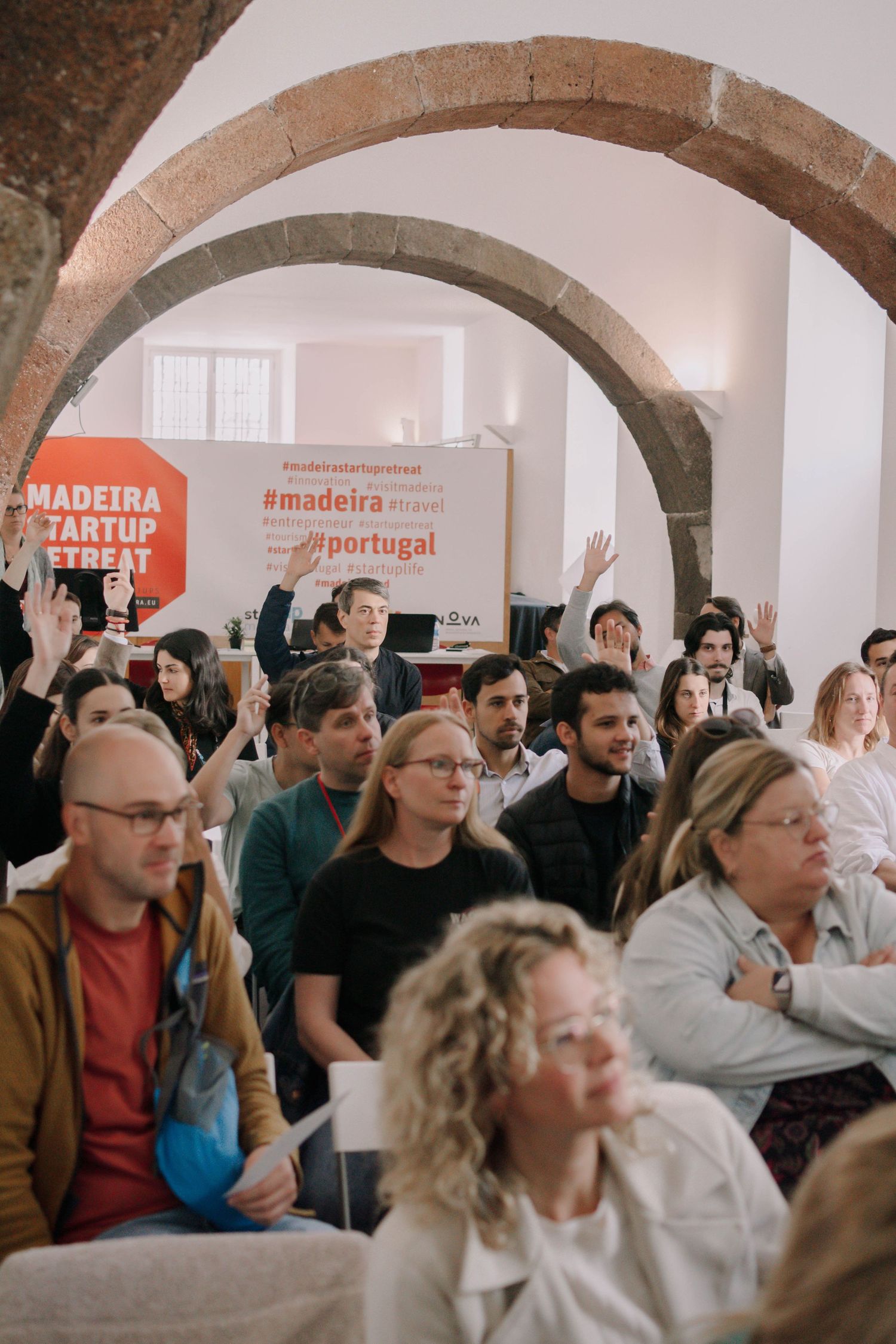 Audience raising hands at Madeira Startup Retreat event in Portugal, seated in a historic arched venue.